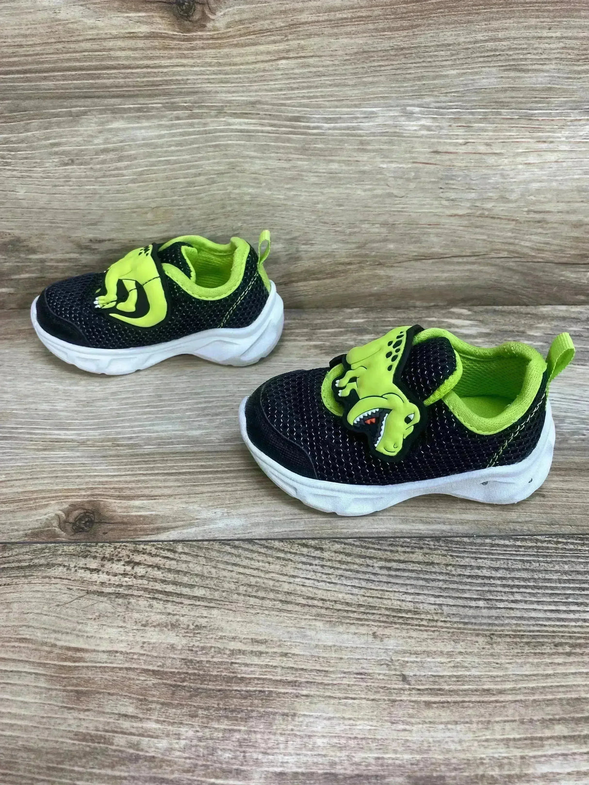 Front-right view of Carter's toddler dinosaur light-up shoe showcasing neon green lining and rubber dinosaur strap with white sole on wood background