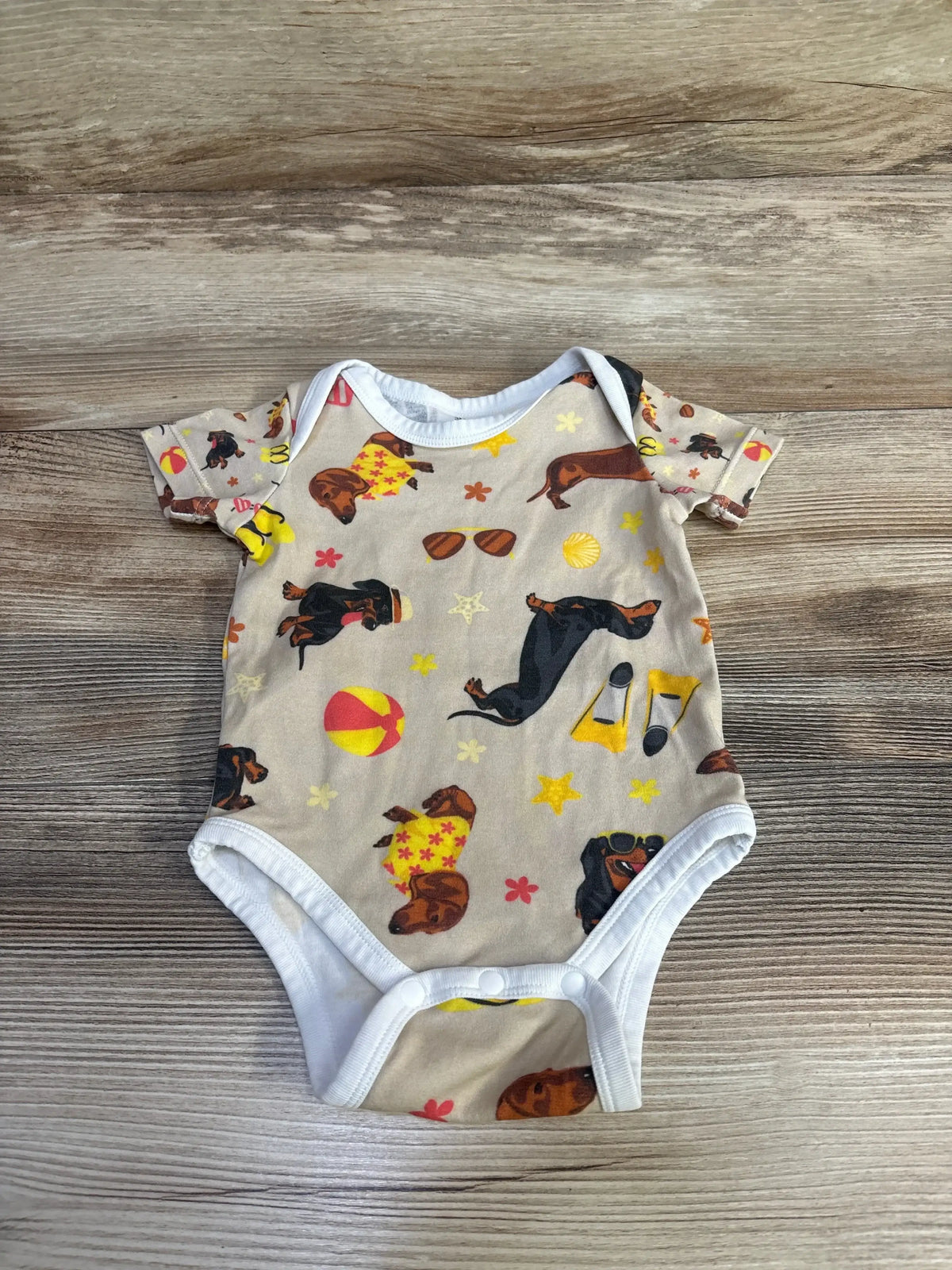 Top-down view of tan dachshund-print baby bodysuit (3M) showing short sleeves, white trim and snap crotch on wood background