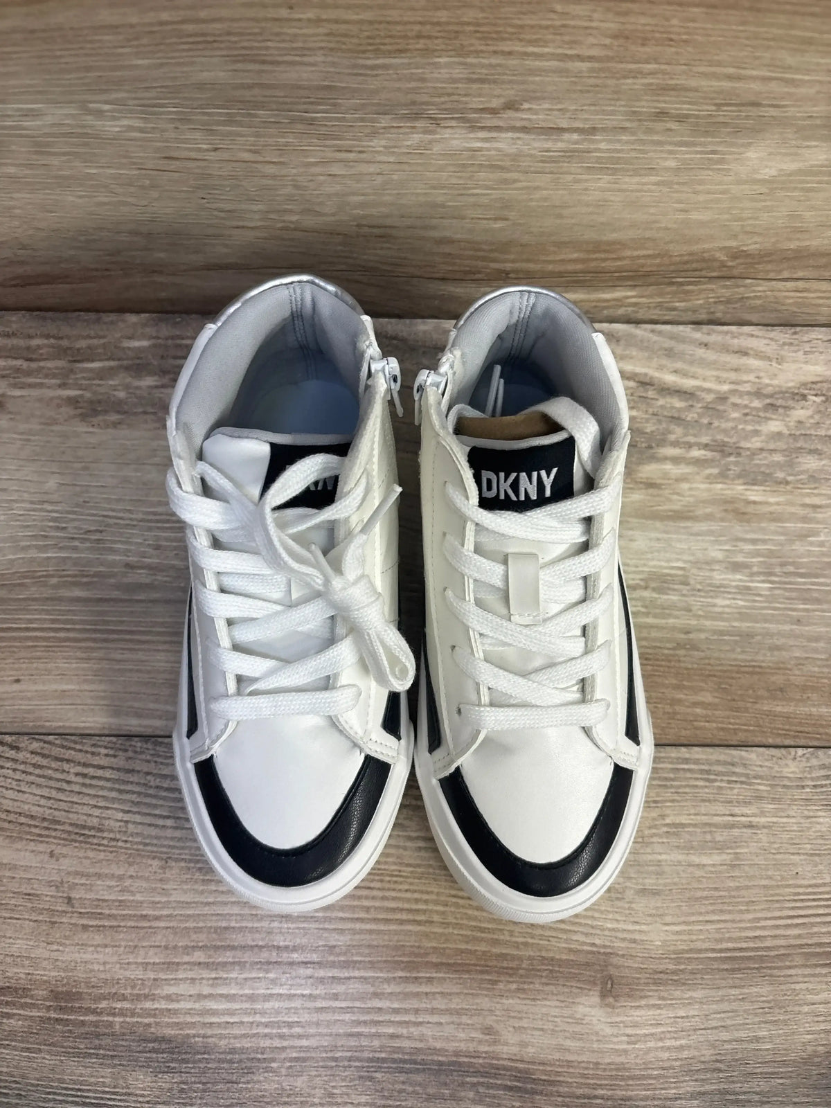 Top-down view of DKNY girls' high-top sneakers, white with black toe accents and DKNY tongue logo on wood floor