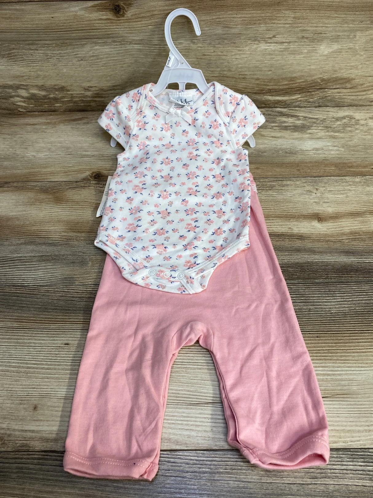 Front view of white floral baby bodysuit with bow paired with pink pants on hanger against wood background