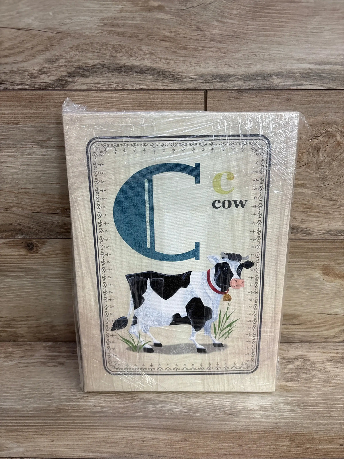 Front view of Oopsy Daisy cow painting on wood panel, large blue letter C and cow illustration, shrink-wrapped on wood floor
