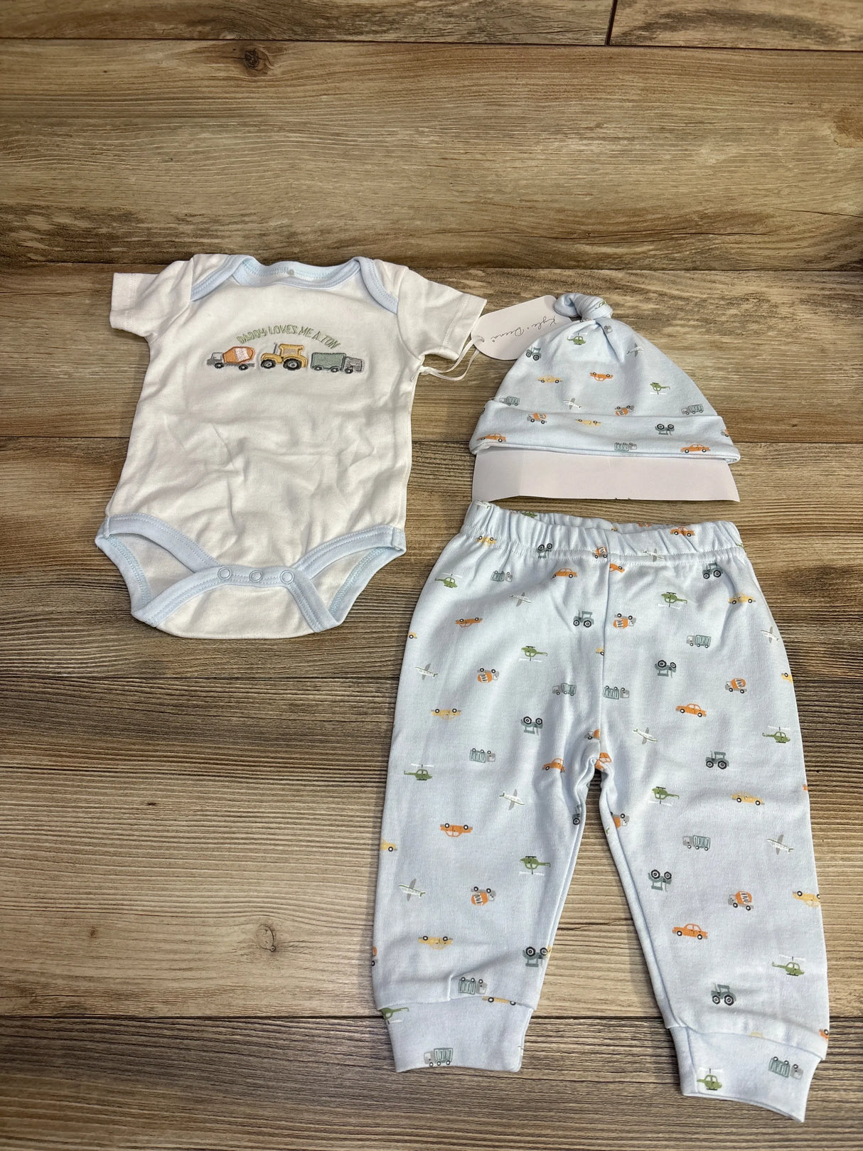 White baby bodysuit with trucks and 'Daddy loves me a ton' text, light blue pants and matching hat with vehicle prints