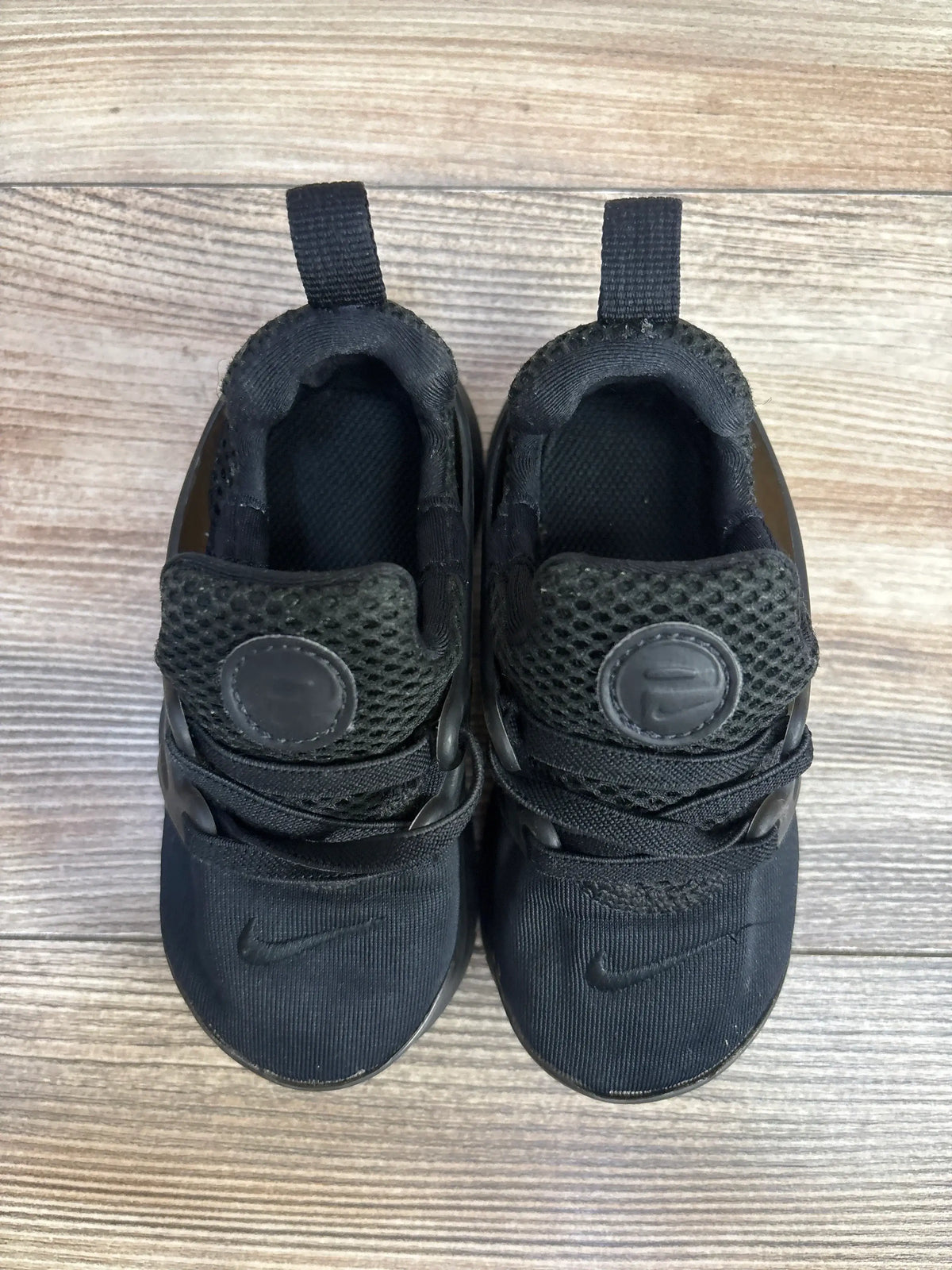 Black Nike toddler sneakers on wooden floor, mesh upper, prominent swoosh logo visible