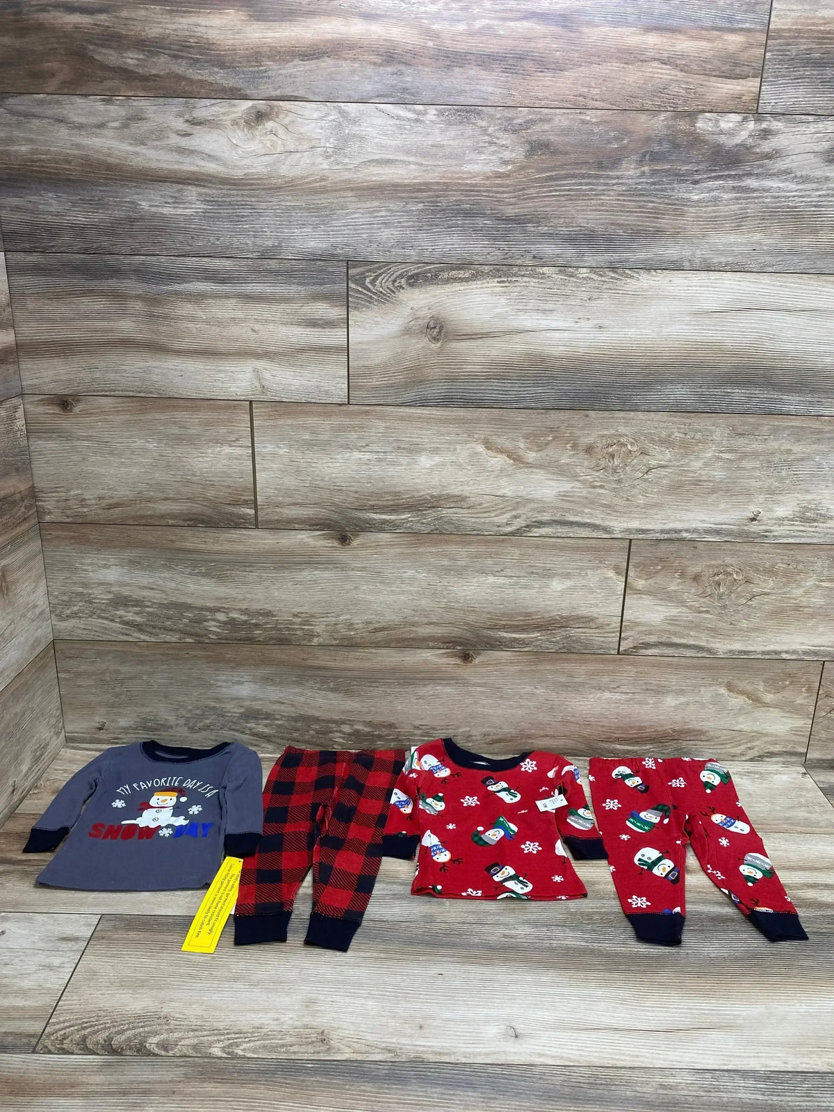 Flat-lay of two 12M toddler pajama sets showing a gray long-sleeve snow day top with buffalo-plaid pants and a red snowman-print top with matching pants on wood plank floor