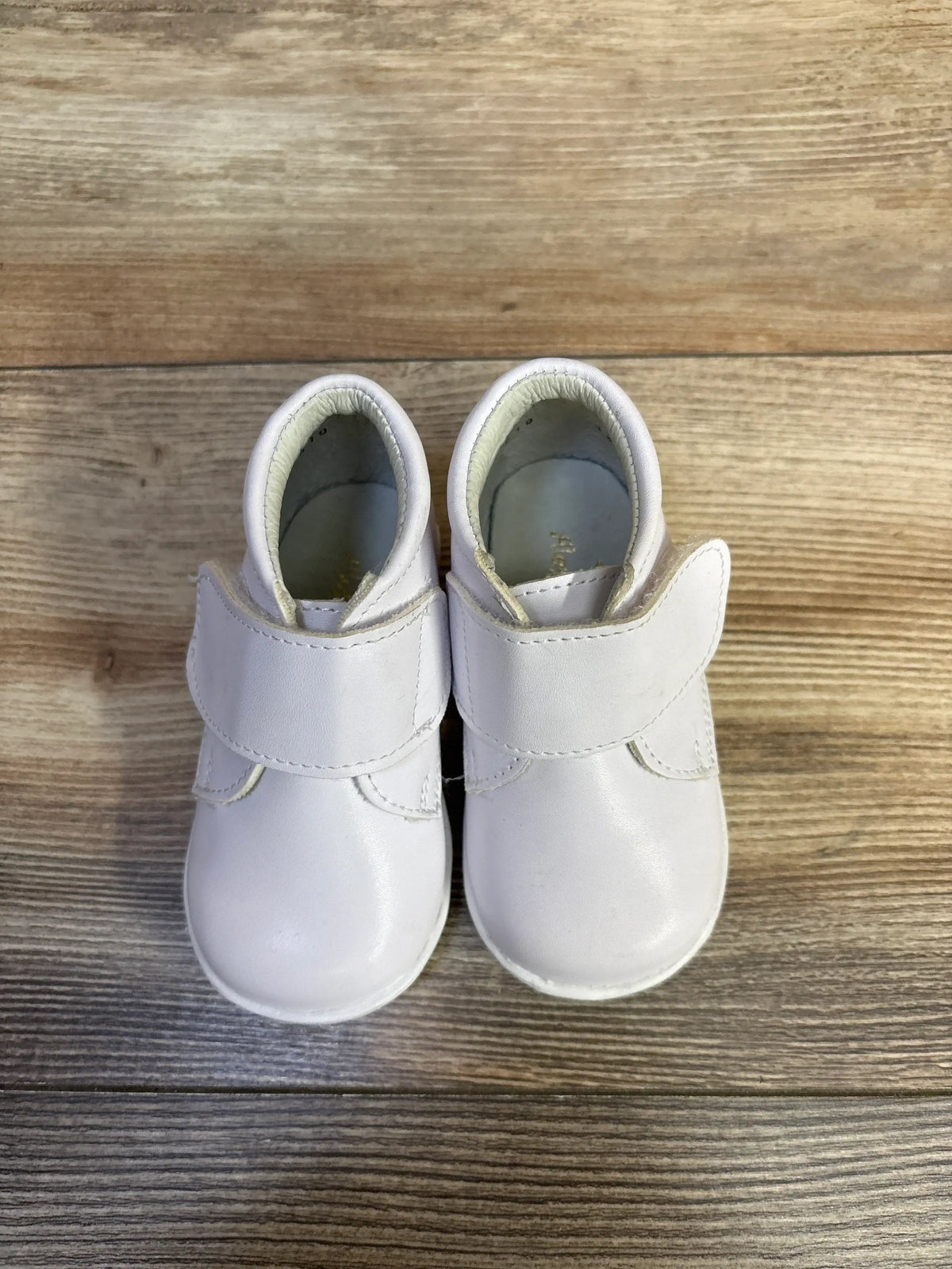 A pair of white Alegria Kids patent leather first walker shoes with velcro straps on a wood background.