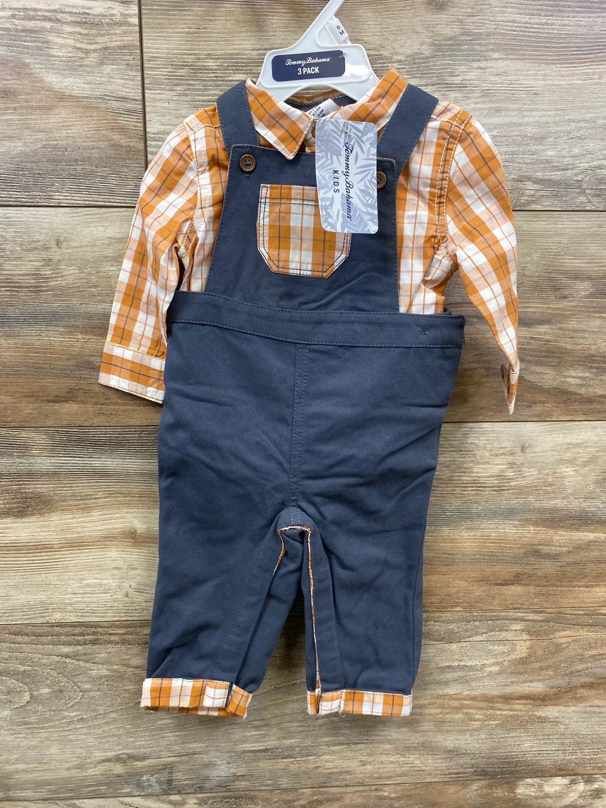 Front view of Tommy Bahama 3-piece baby set: orange plaid shirt and navy overalls with chest pocket and brand tag on wood