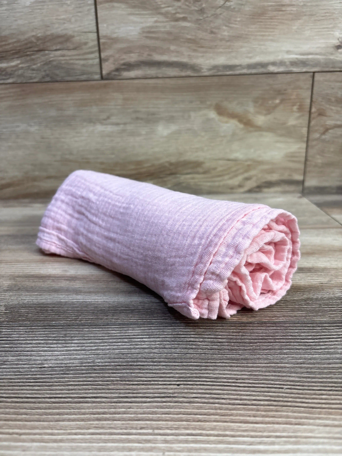 Rolled pink Little Me muslin swaddle blanket, 36 by 40 inches, showing its textured fabric on a wood-look floor