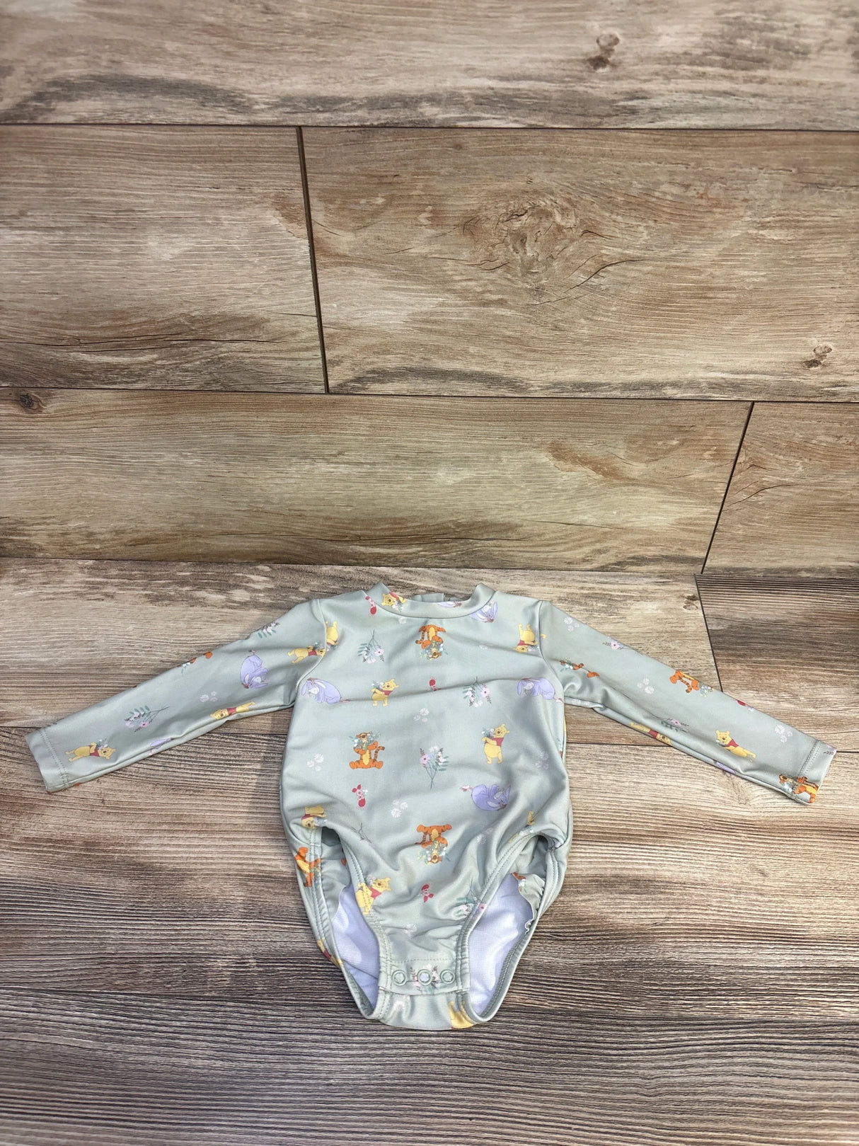 Flat lay of green Winnie the Pooh rashguard swimsuit size 9M with snap crotch and all over print on wood floor