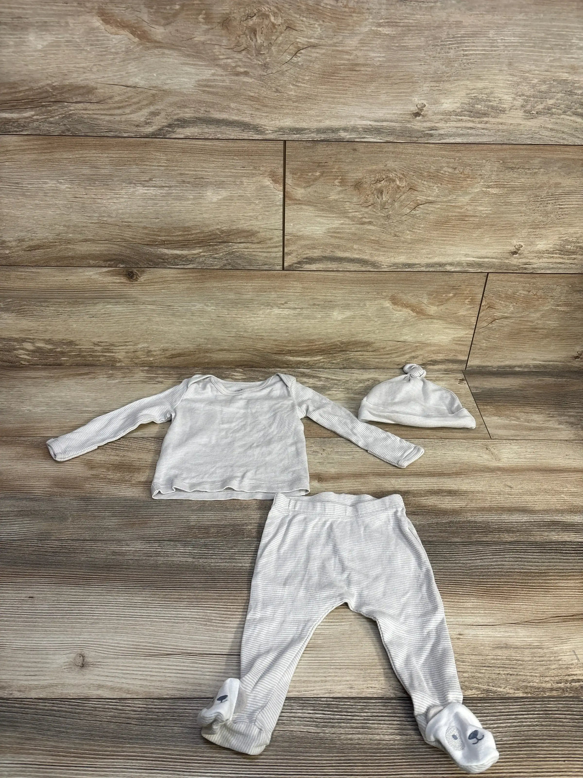Flat lay of beige baby 3 piece set: long sleeve top, footed pants with animal faces, and knotted hat on wood floor