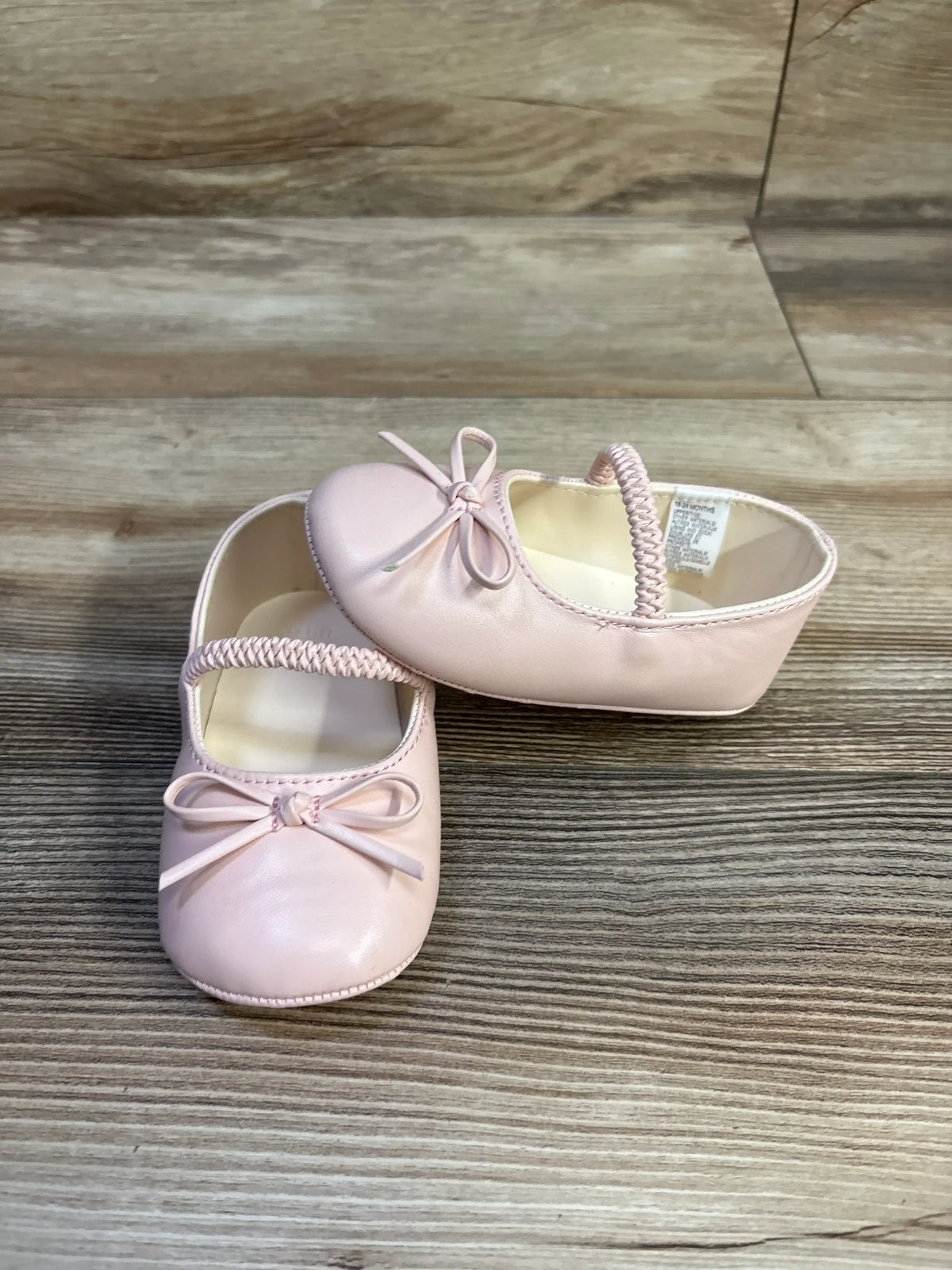 Angled view of Janie & Jack pink soft sole baby ballet flats showcasing braided elastic strap and bow detail on woodgrain floor