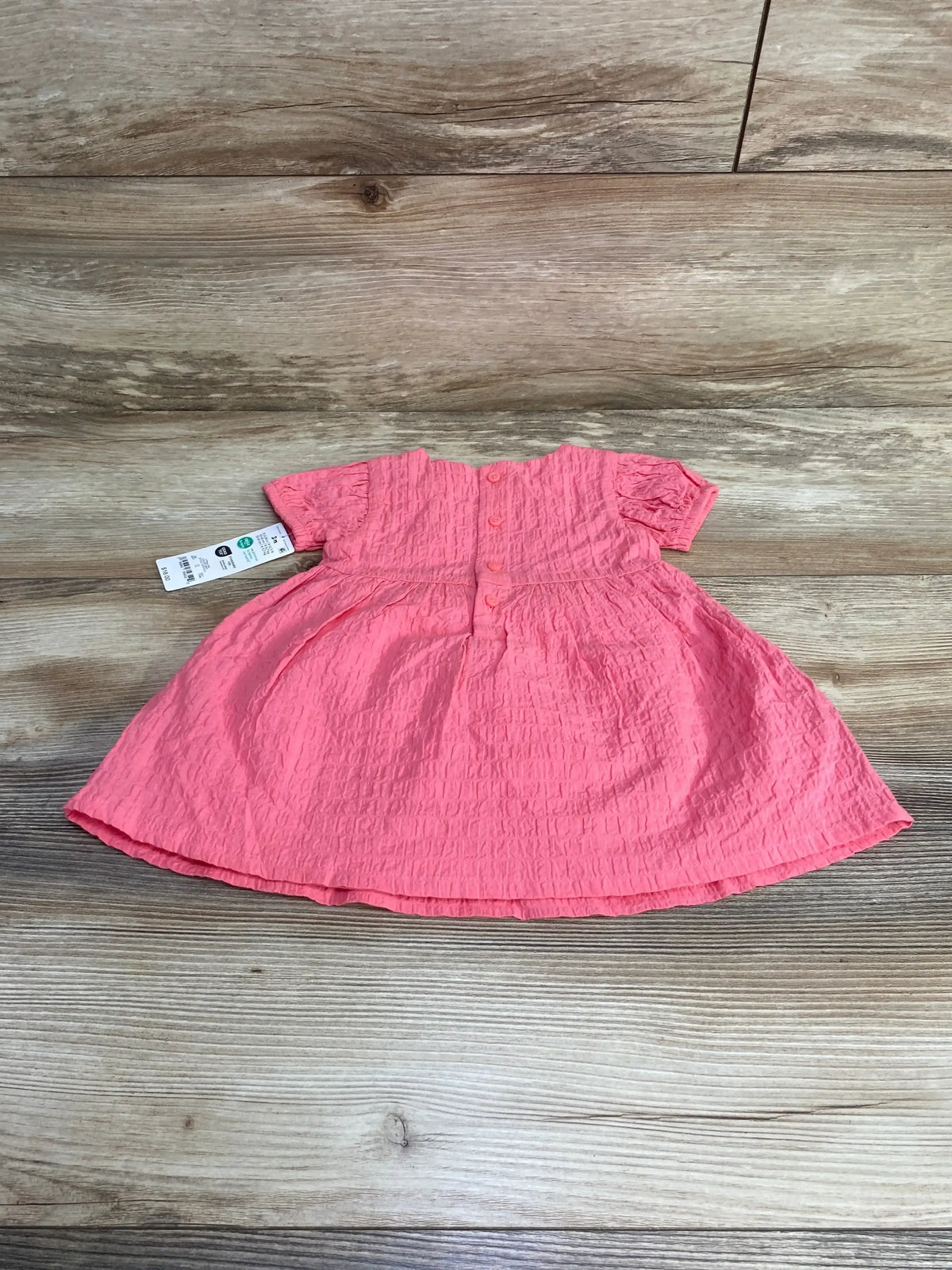 Flat lay of pink 3M baby dress showing back button closure, crinkled textured fabric and short puff sleeves on wood plank background