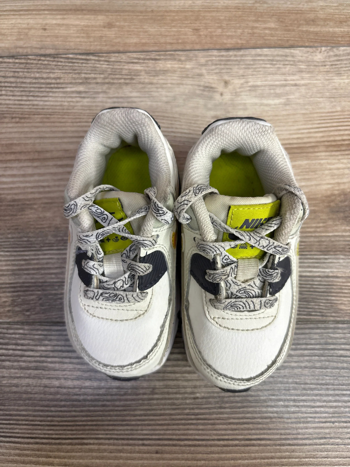 Pair of white Nike toddler sneakers with black accents and patterned laces on wooden floor