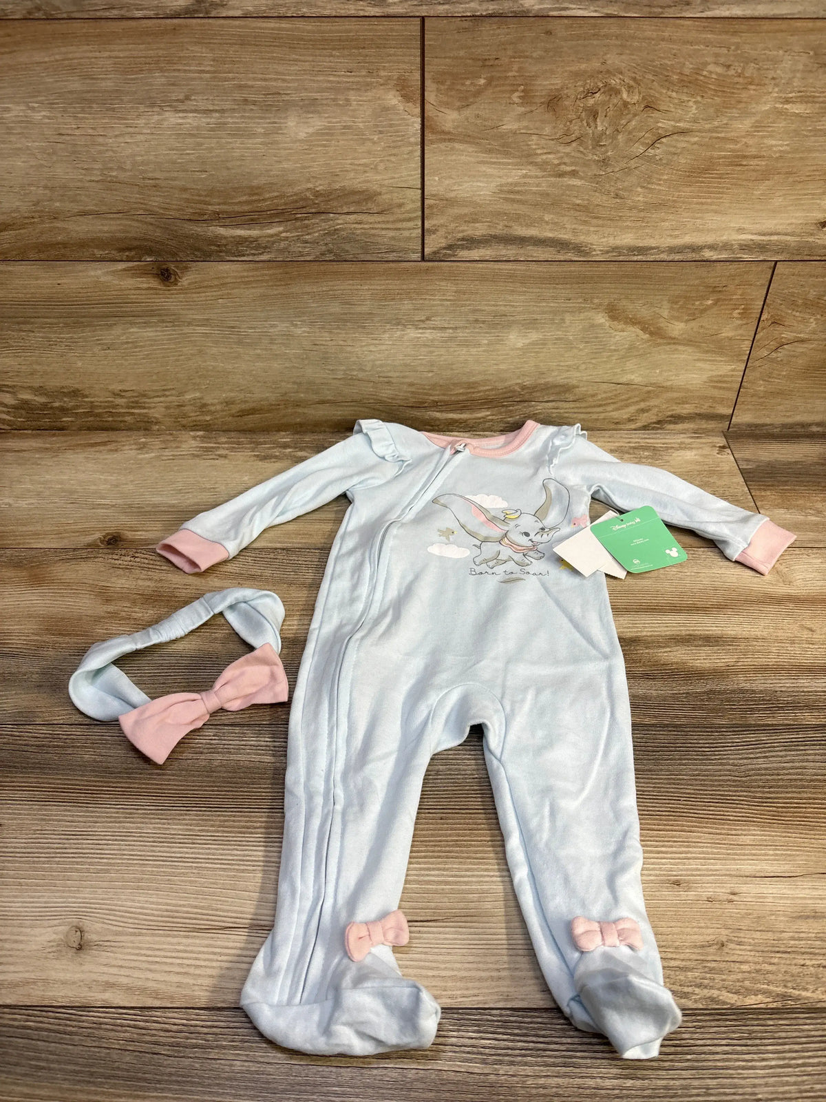 Flat lay of Disney Baby Dumbo footed sleeper and matching headband showing Dumbo print, pink trim, and foot bows on wood background