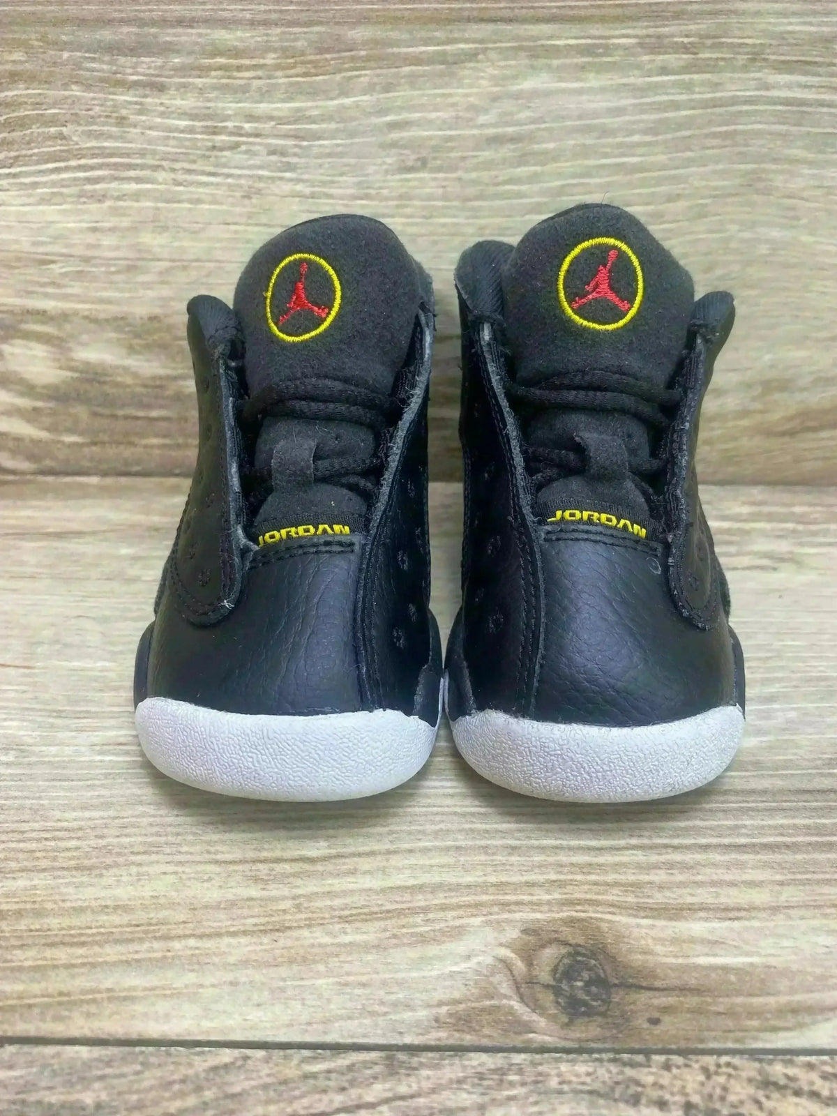 Front view of Air Jordan 13 Retro toddler sneakers showing black leather and suede, red Jumpman on tongue, white soles on wood