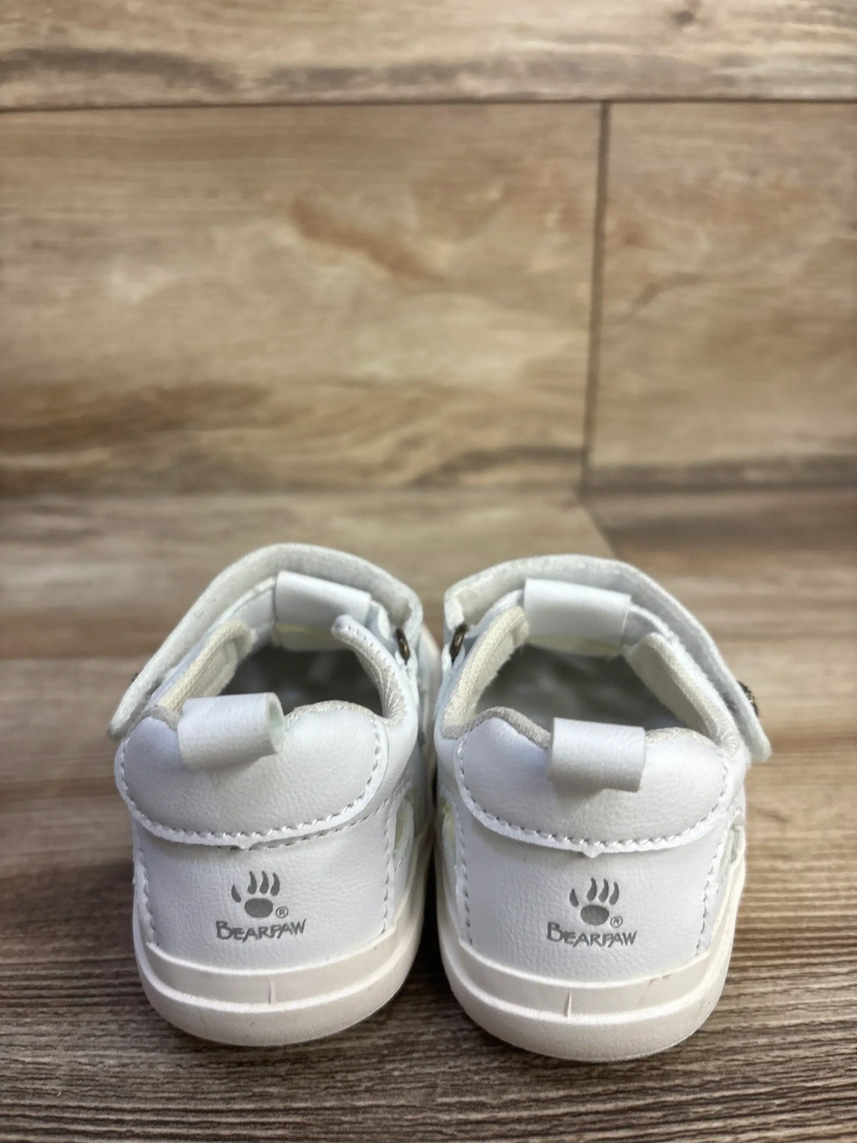 Rear view of white Bearpaw toddler fisherman sandals showing heel pull tabs, Velcro straps, Bearpaw logo on wood floor background