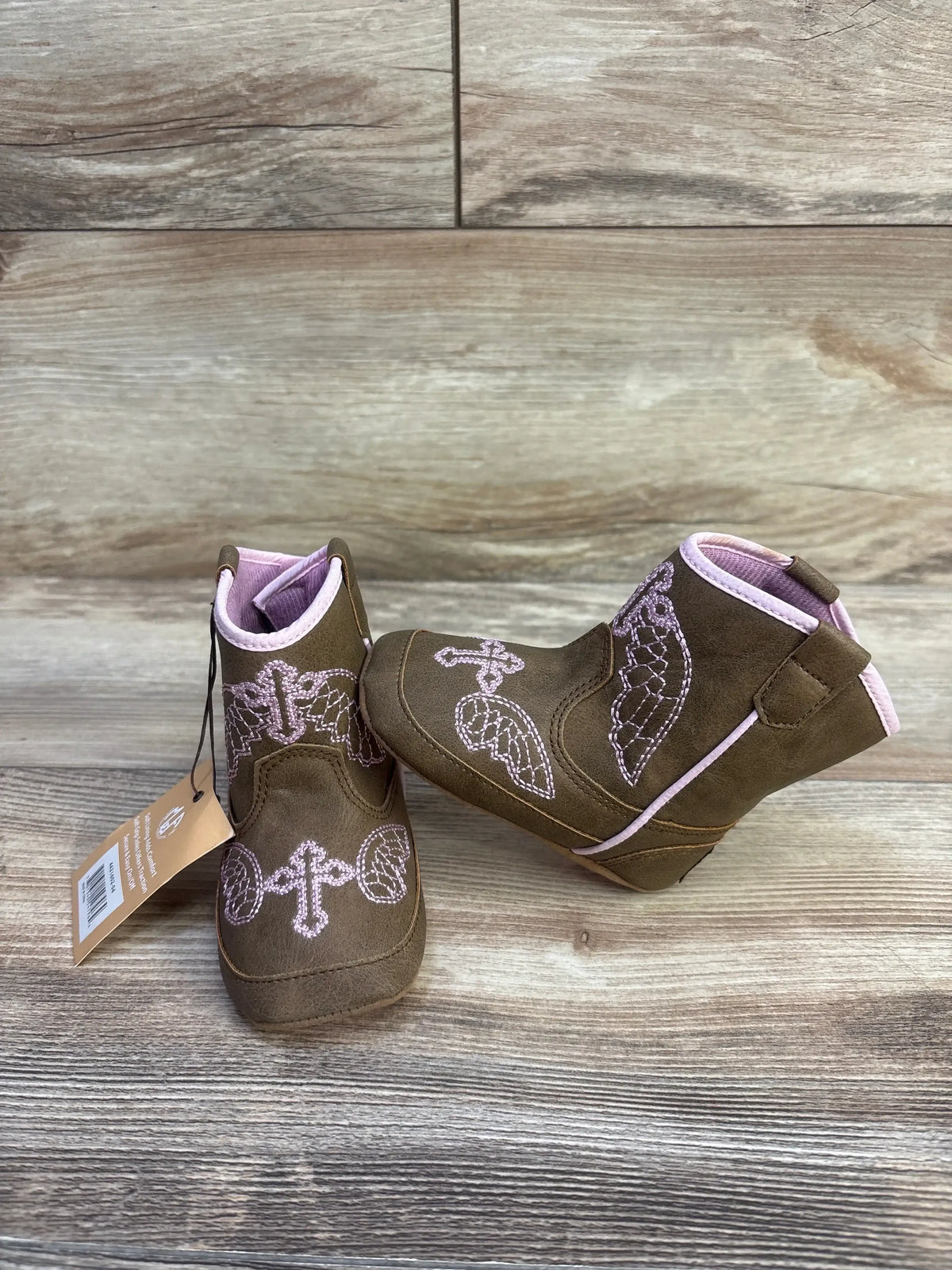Front and side view of Twister Gracie baby buckers boots in brown with pink embroidered crosses and wing motifs on a wood background