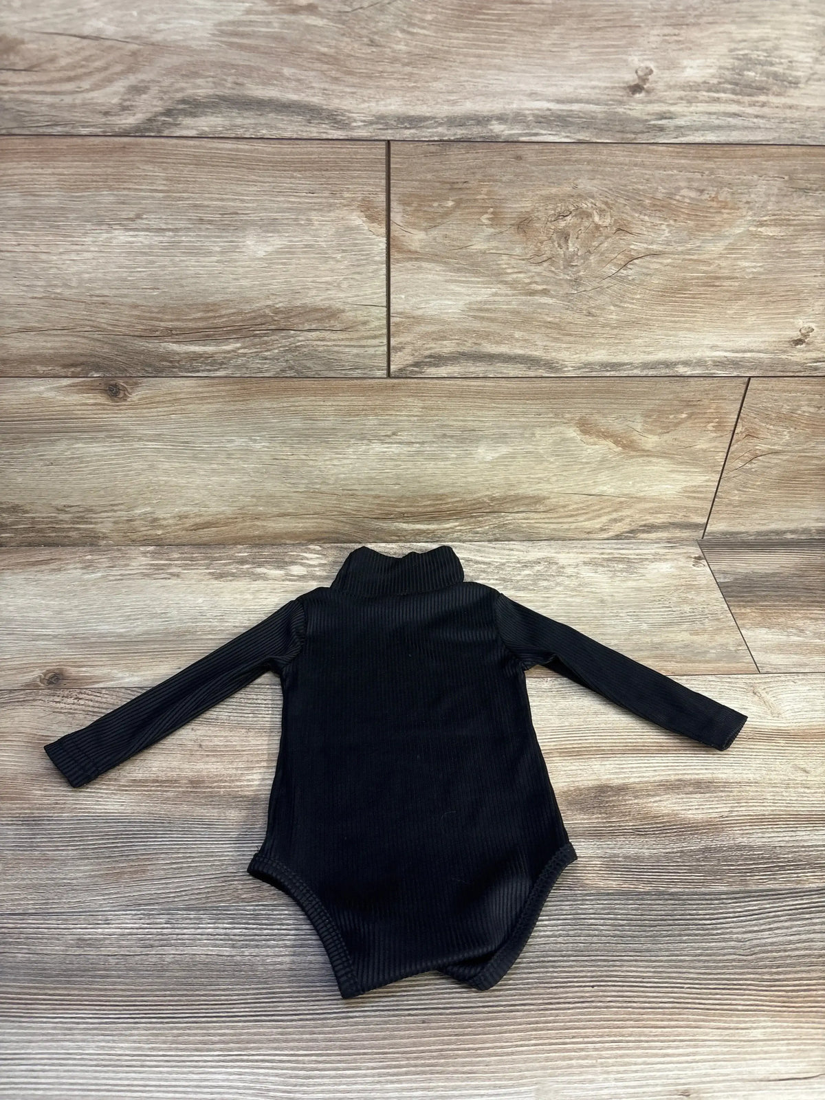 Top-down of black ribbed turtleneck baby bodysuit showing ribbed fabric and three snap crotch on wood floor