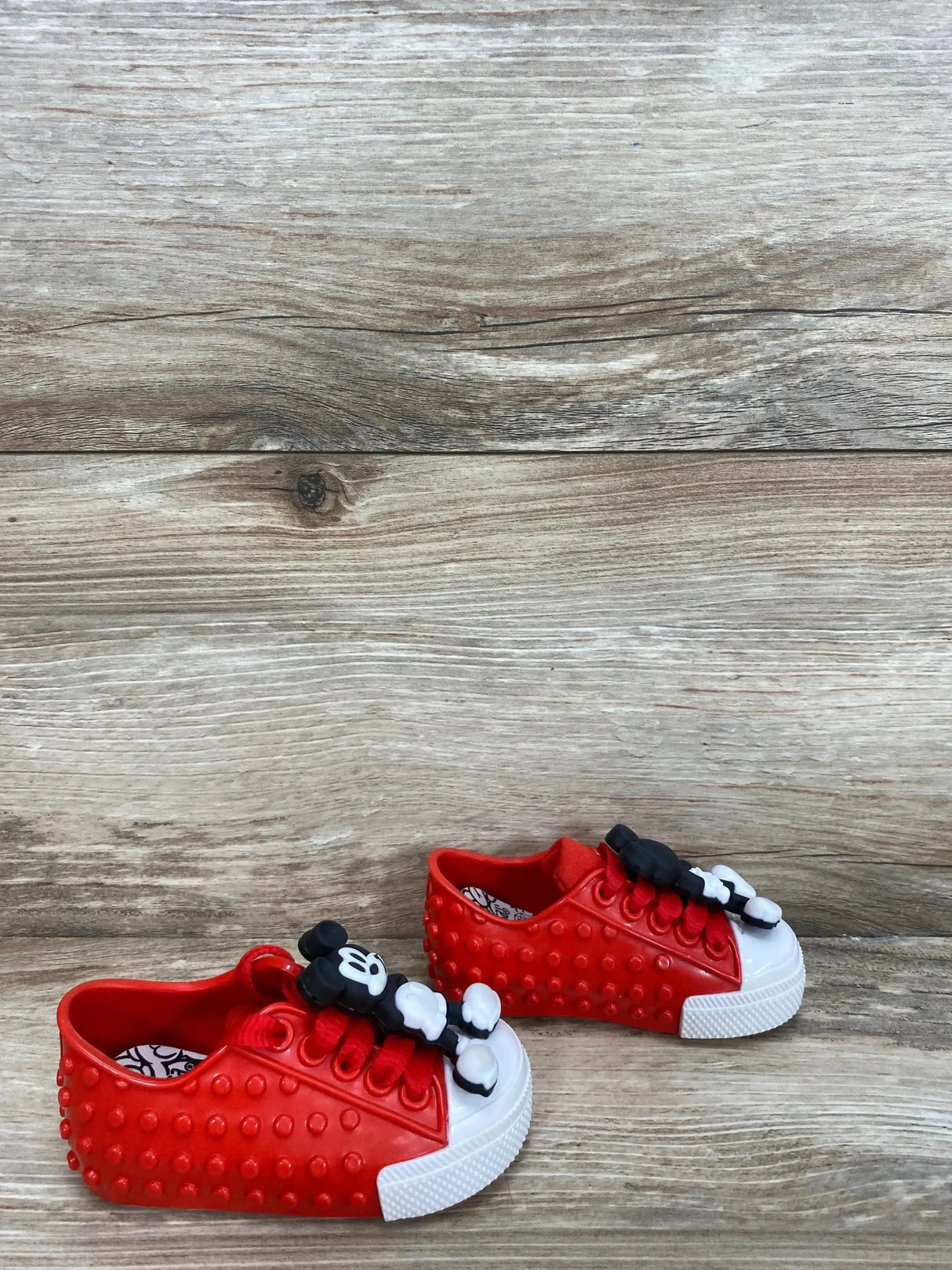 Angled view of red Mini Melissa Polibolha sneaker with raised studs, white toe cap and Mickey bow on laces on wood backdrop