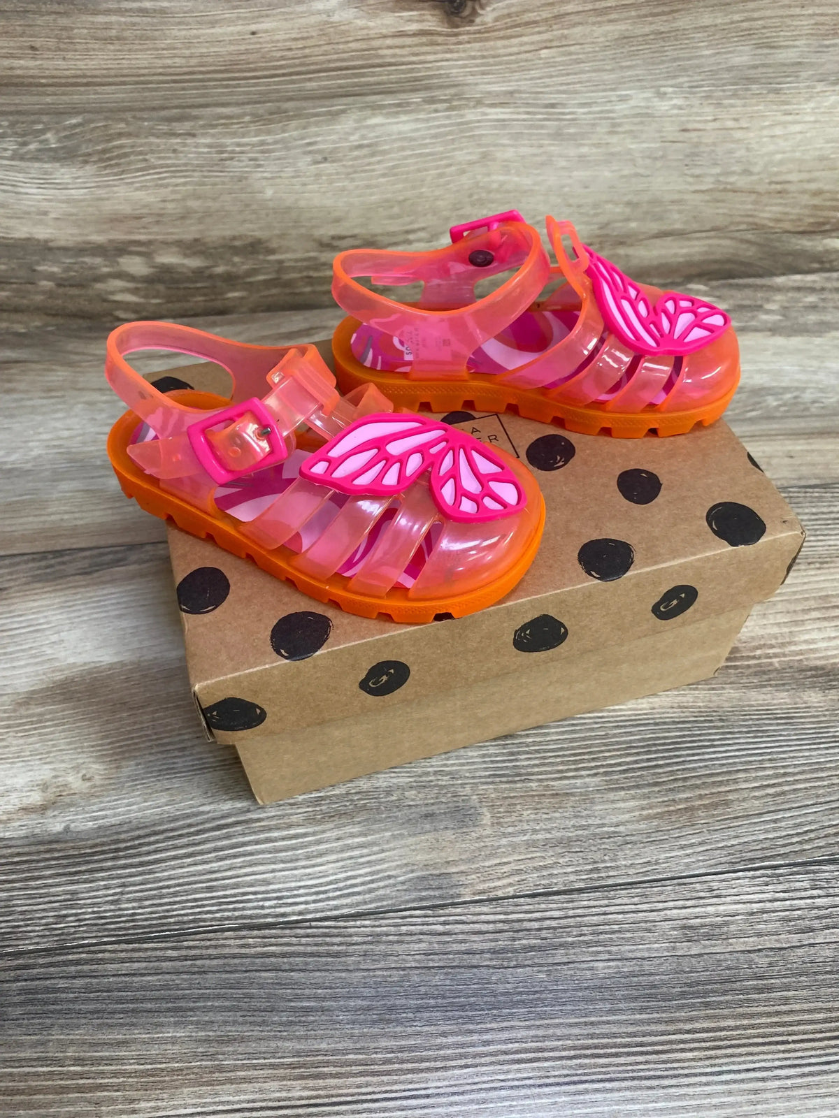 Three-quarter view of Sophia Webster toddler jelly sandals showcasing pink butterfly appliques and orange soles on a polka-dot shoebox background
