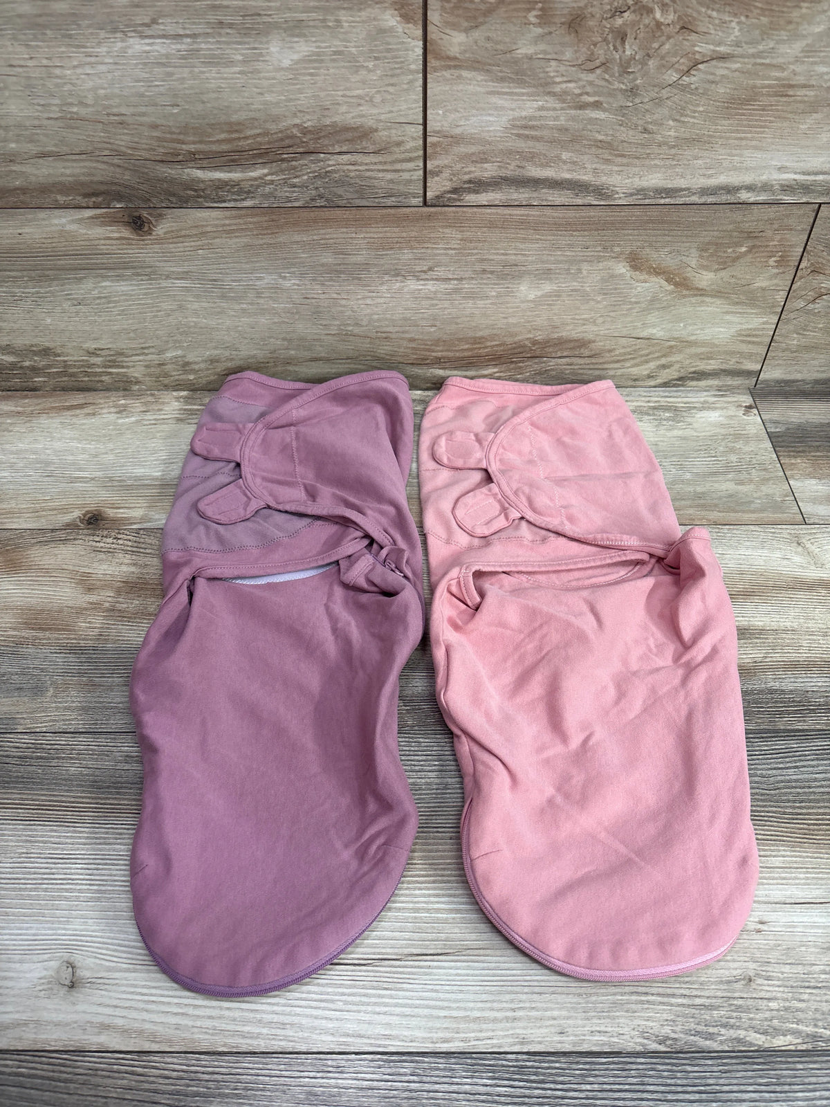 Top view of two baby swaddle wraps in mauve and pink showcasing wrap closures on a wood-grain background