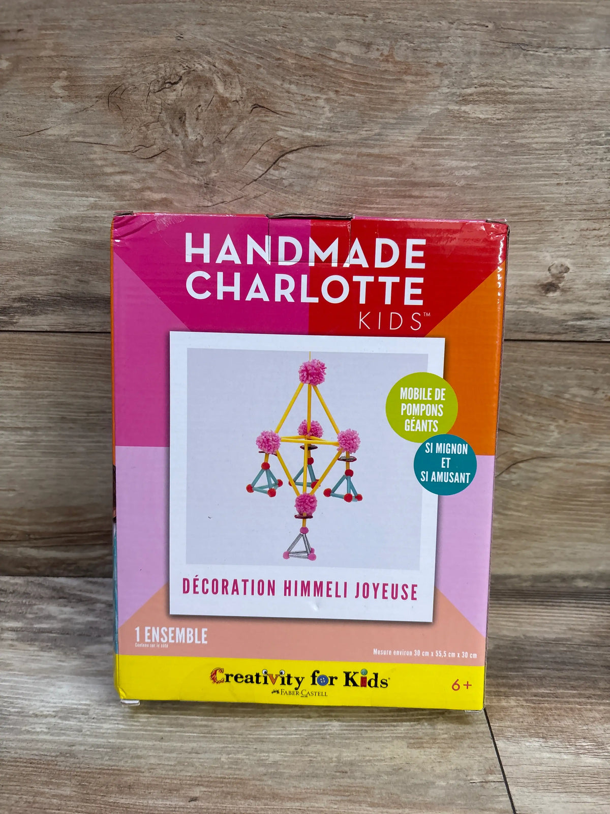 Front view of Handmade Charlotte Kids Happy Himmeli Craft Kit showing a colorful pom-pom mobile on a wood floor