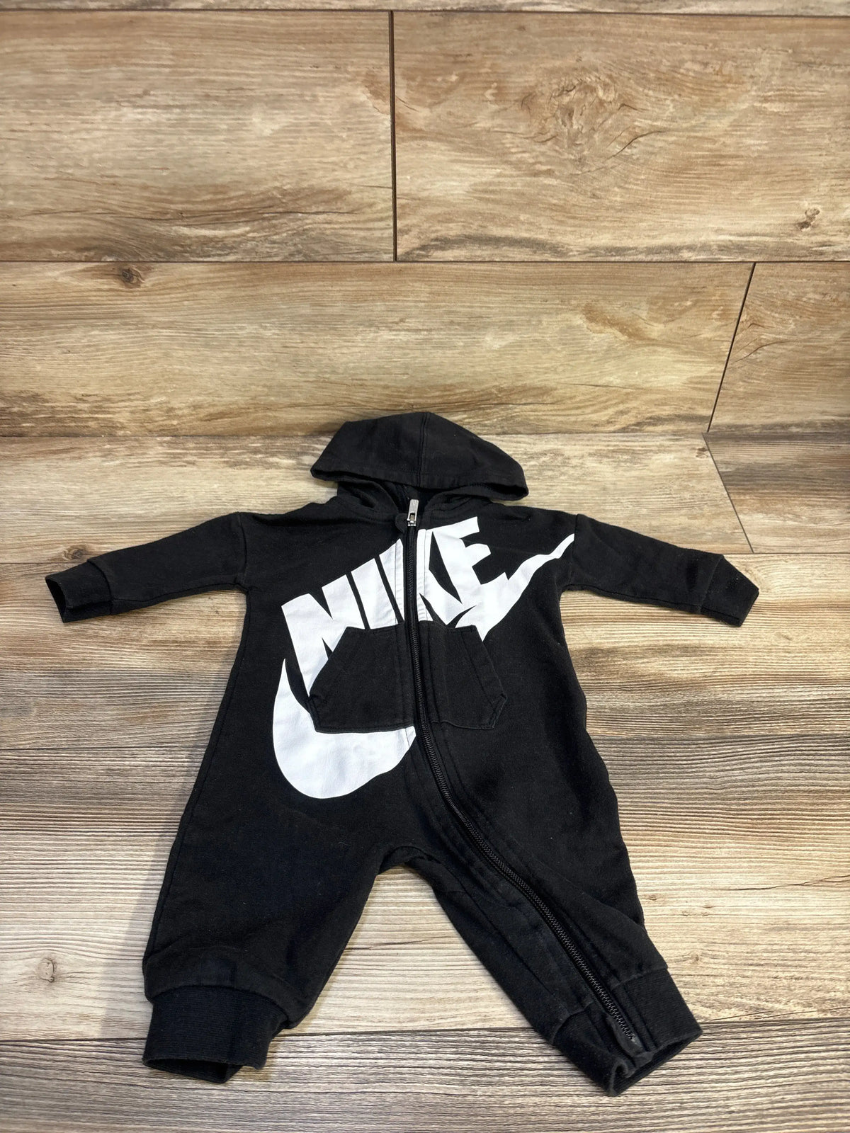 Front view of Nike black hooded coverall showcasing white Nike logo, front zipper, and hood on wood-look flooring