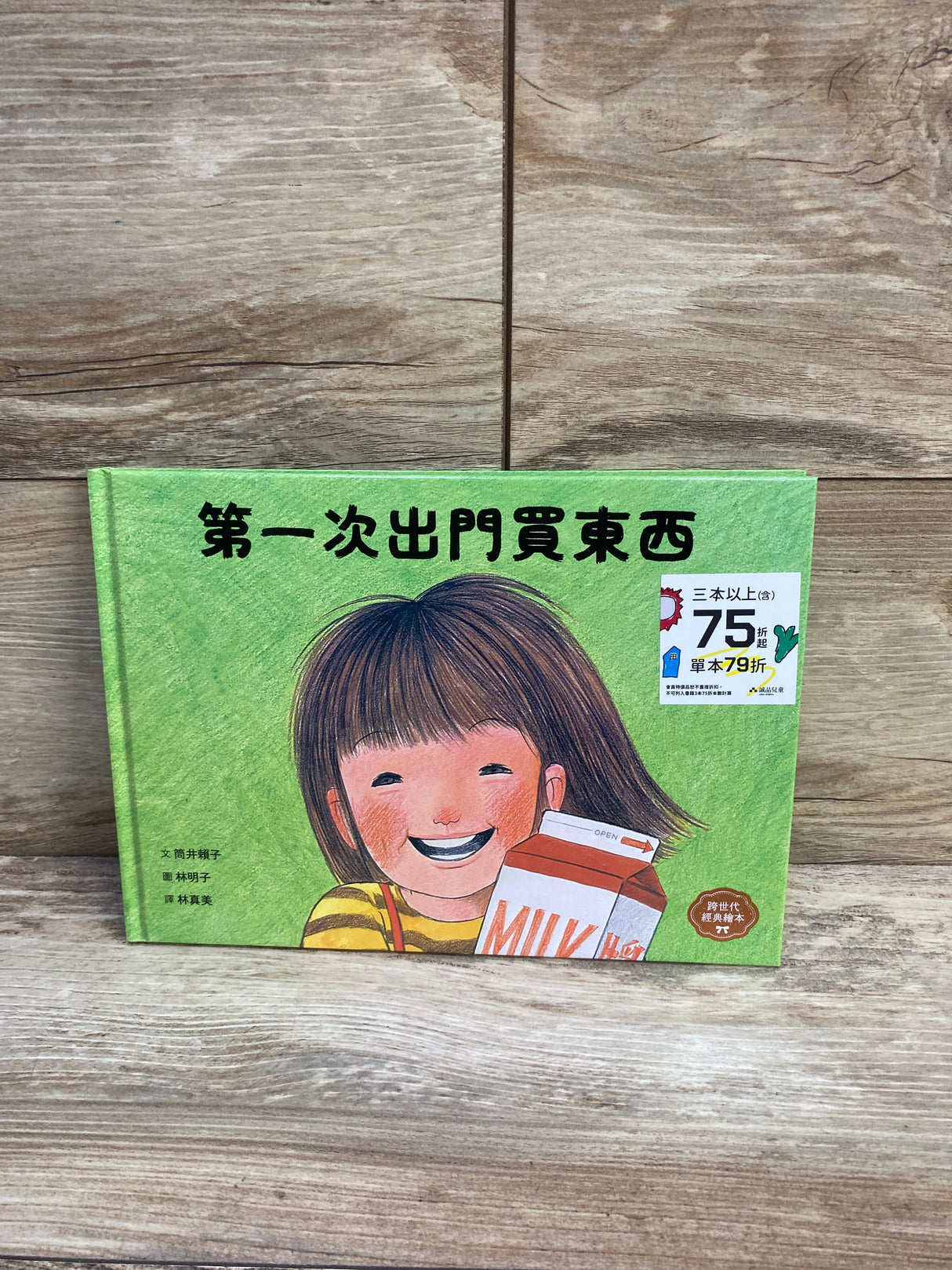 Front cover of green hardcover kids picture book ‘第一次出門買東西’ showing a smiling child with a milk carton on wood