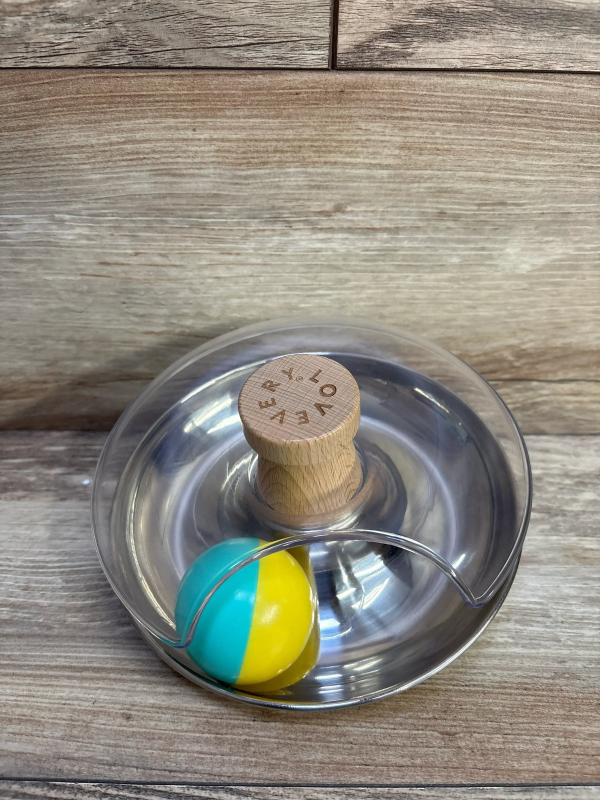 Top view of Lovevery Tip & Turn toy with stainless steel spinning base, clear cover, wooden knob engraved LOVEVERY, and teal-and-yellow ball on wood surface