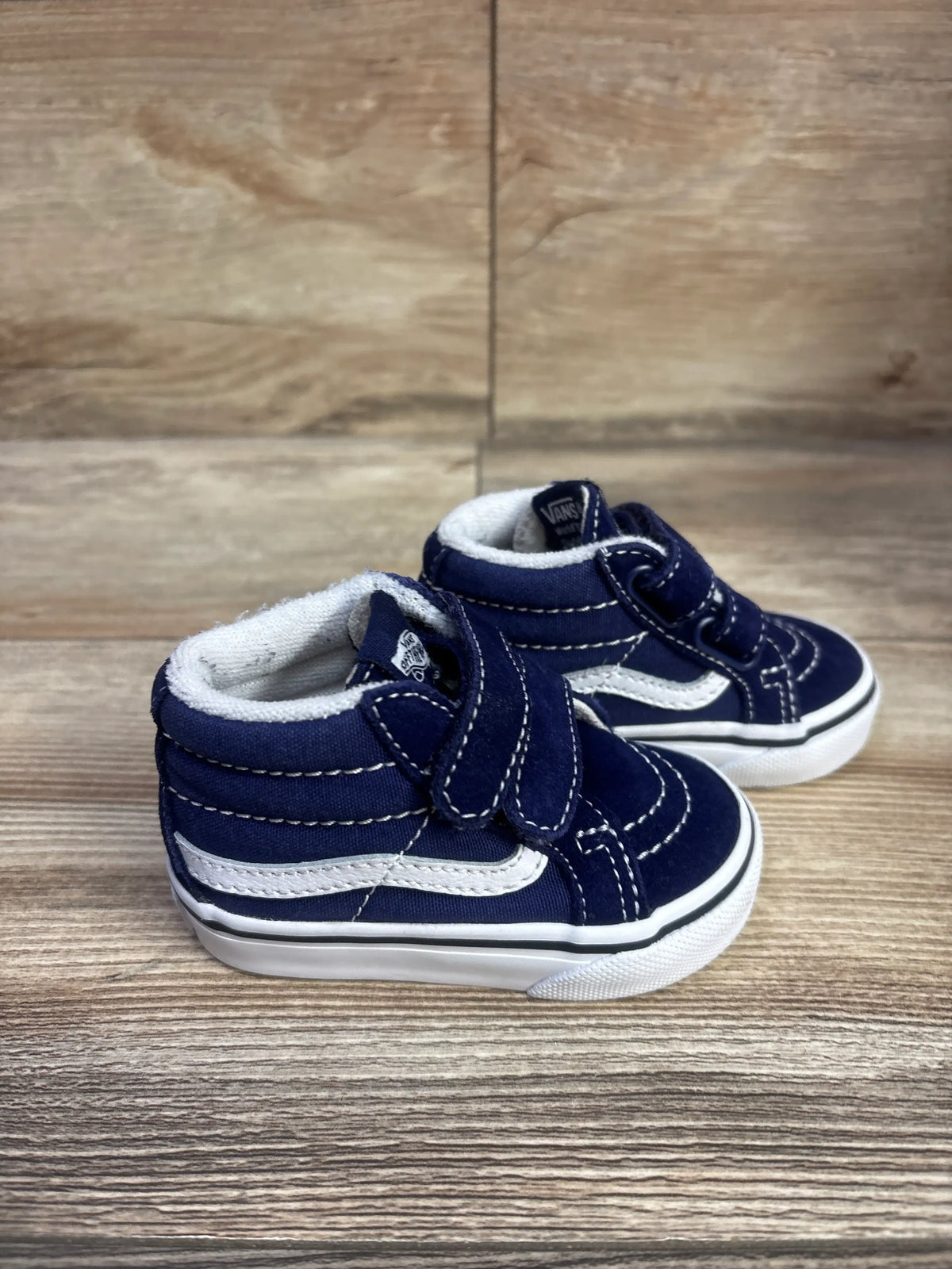 Three-quarter view of navy Vans SK8-Mid V sneakers showcasing white side stripes and hook-and-loop straps on a wood surface