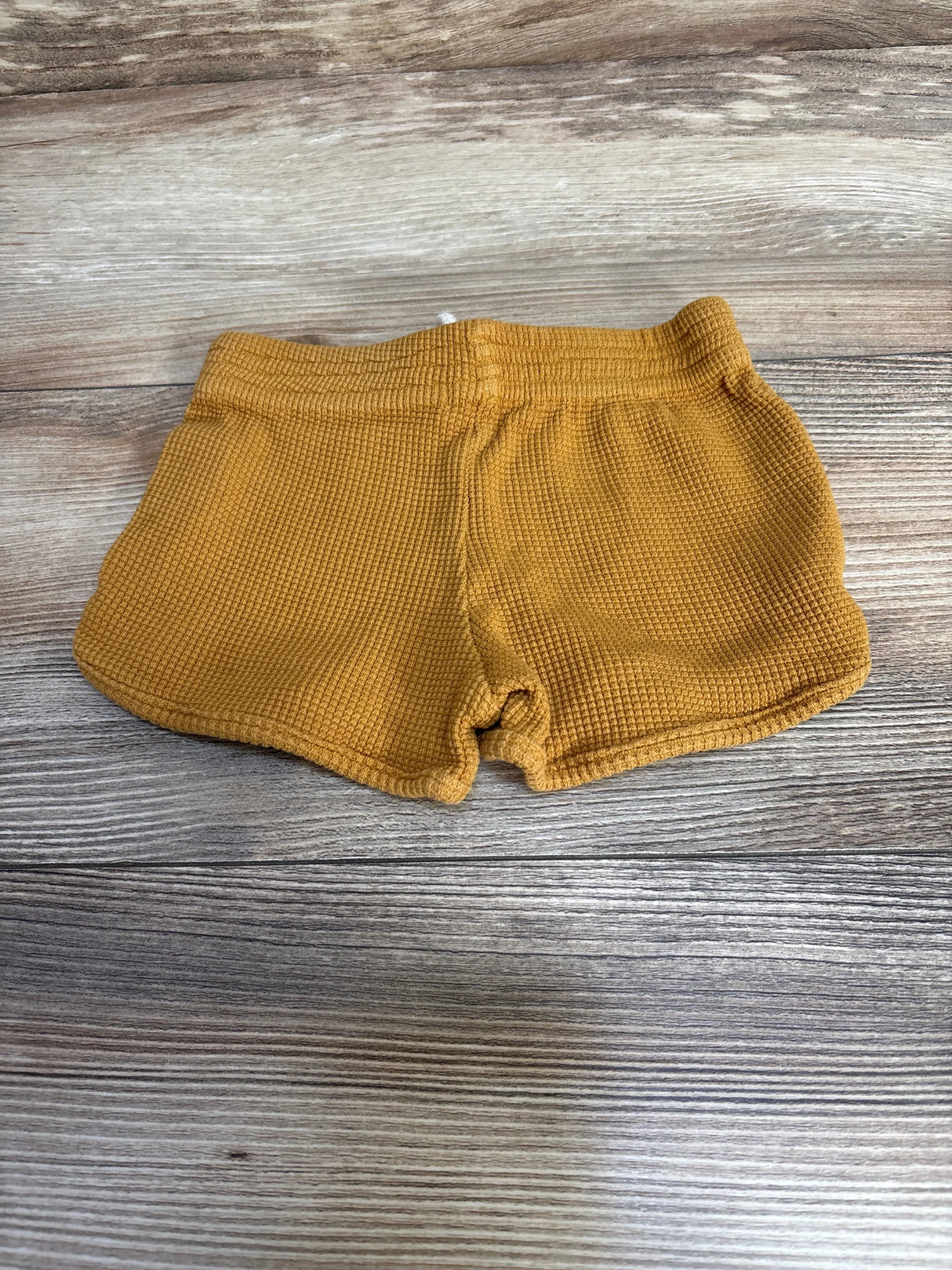 Top-down view of My Little Cozmo organic waffle shorts, mustard yellow, showing waffle texture and elastic waistband on wood background