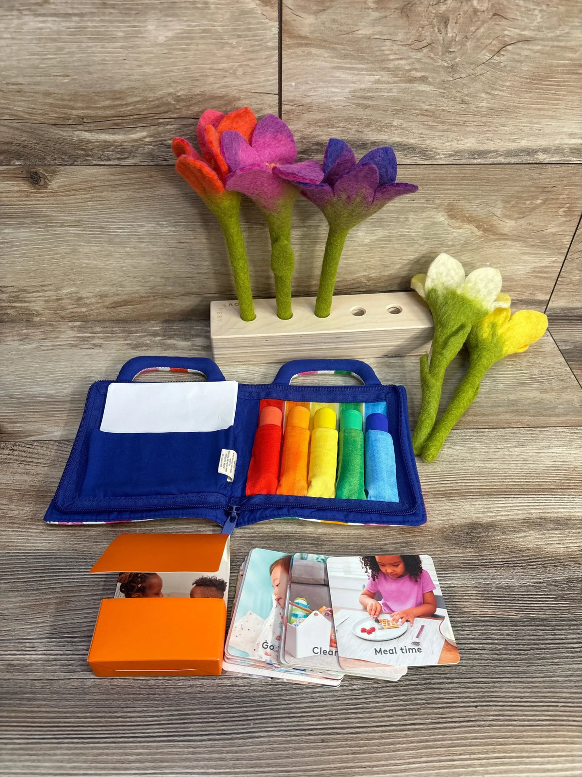Front view of Lovevery The Helper Play Kit with felt flowers in a wooden block, rainbow fabric tubes in a blue zip pouch, and activity cards