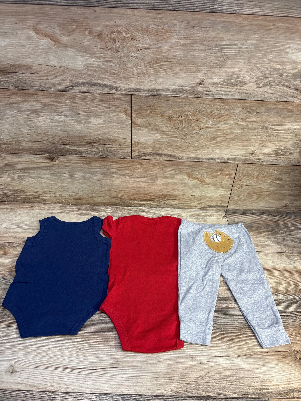 Top view of Carter’s three-piece Home Run set showcasing navy and red bodysuits and gray baseball pants on wood flooring