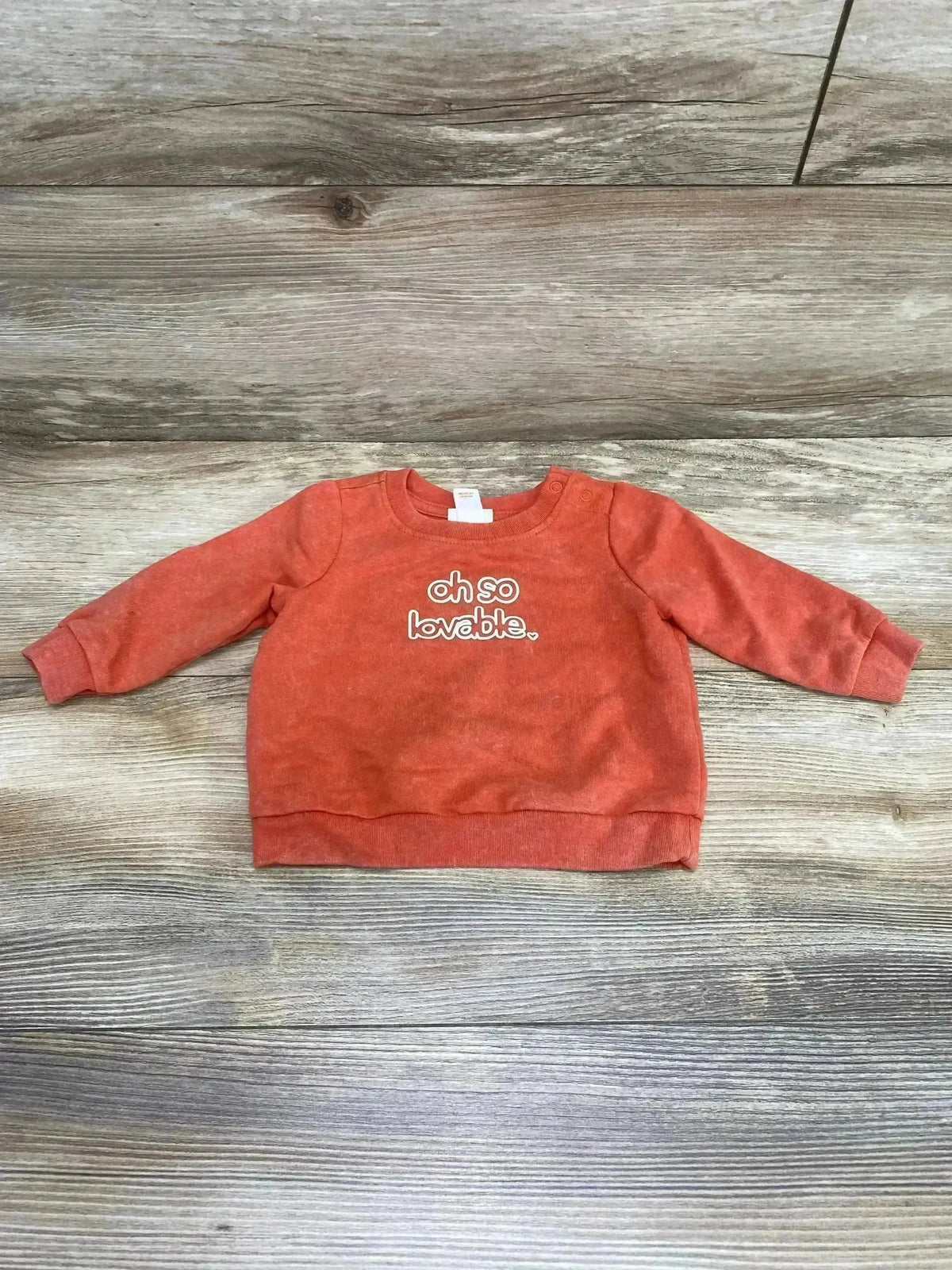 Flat lay of orange Cat & Jack baby sweatshirt size 0-3 months showcasing oh so lovable chest text on wood floor background
