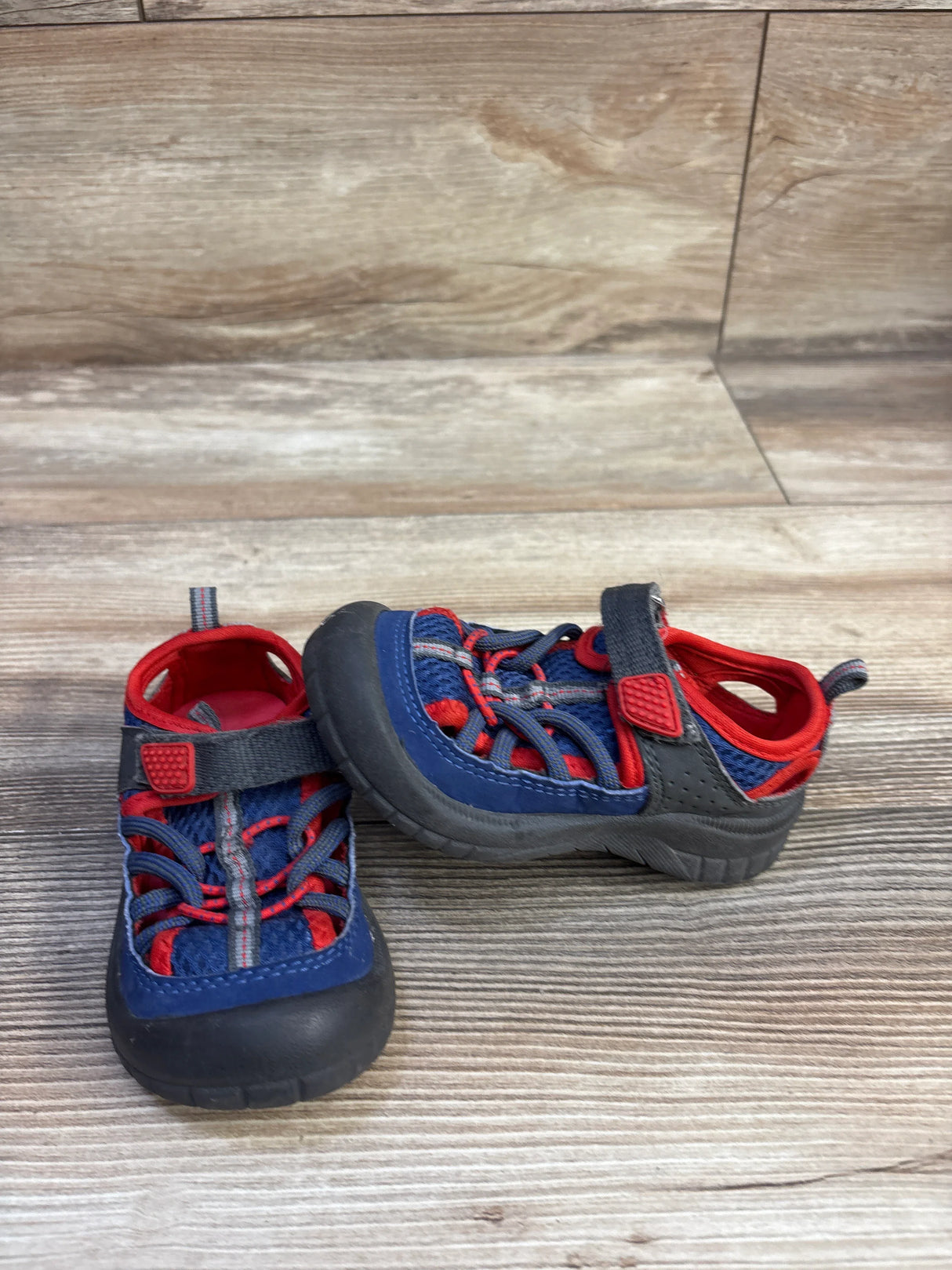 Angled front view of navy and red Oshkosh sandals with rubber toe caps, bungee laces and Velcro strap on wood floor
