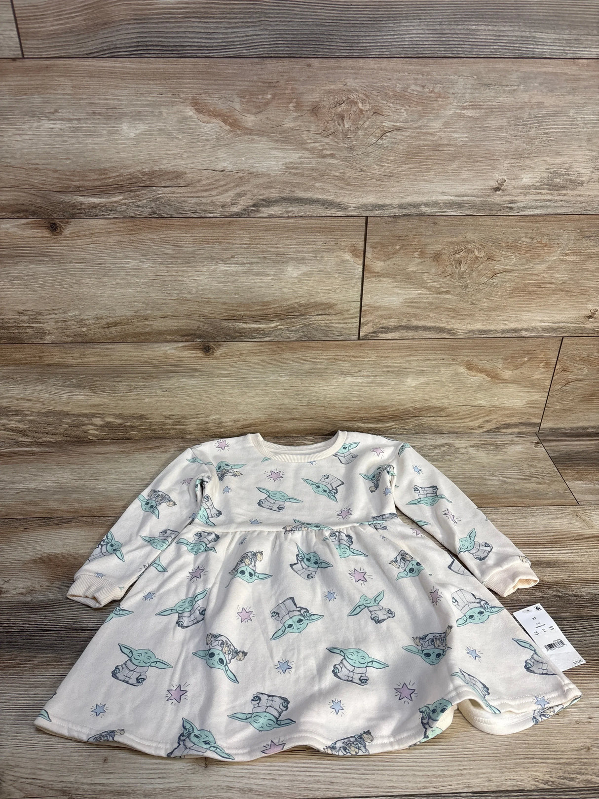 Flat lay of long sleeve baby dress showcasing white fabric with Baby Yoda Grogu print and hangtag on wood plank floor