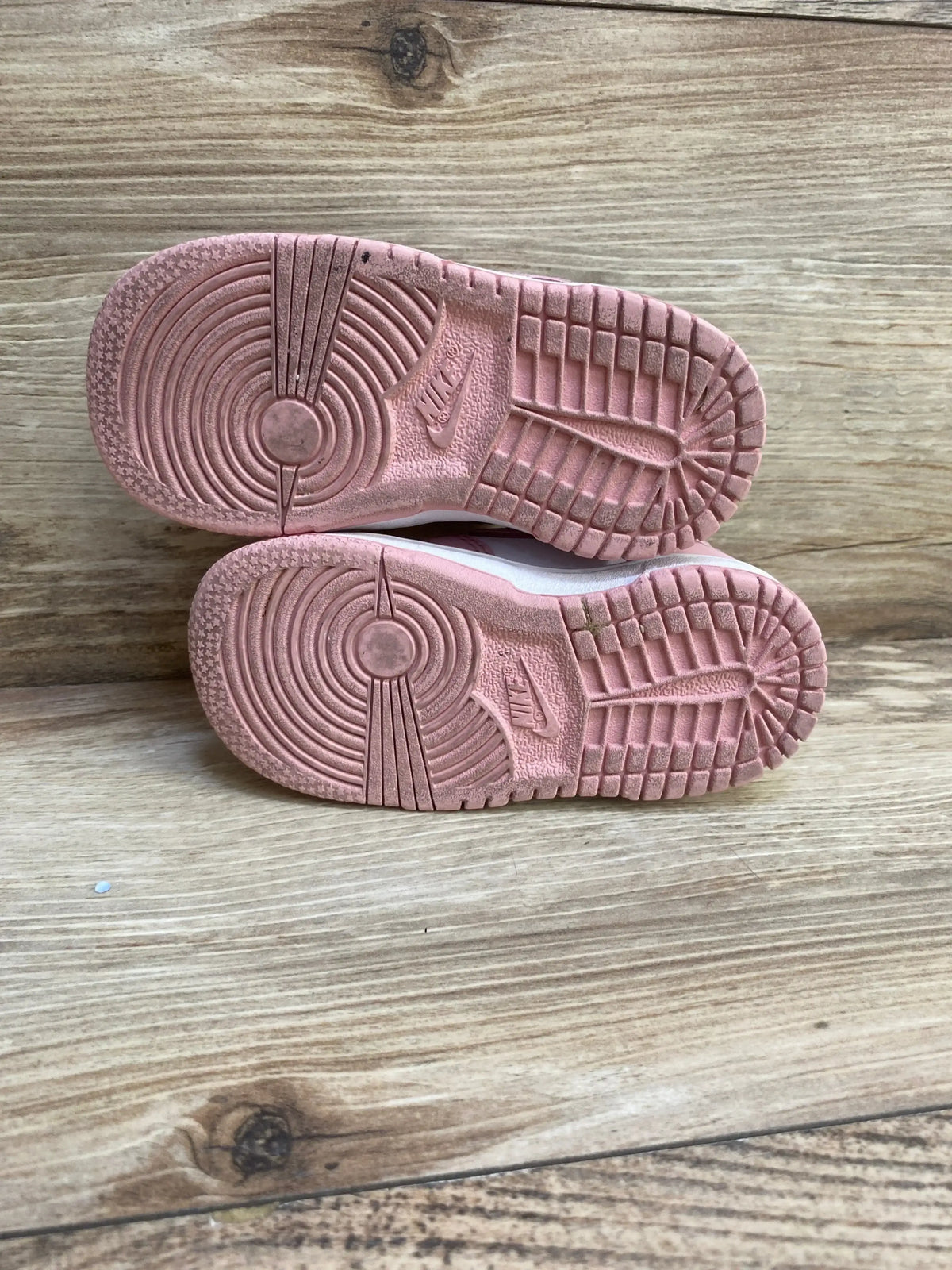 Bottom view of Nike Dunk Low TD ‘Pink Glaze’ toddler sneakers showing pink treaded soles with light wear on wood background