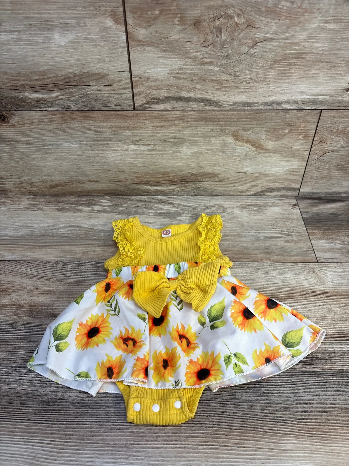 Flat lay of WIBACKER yellow floral ribbed bodysuit dress, size 12 months, with lace trim, bow and snaps on wood flooring