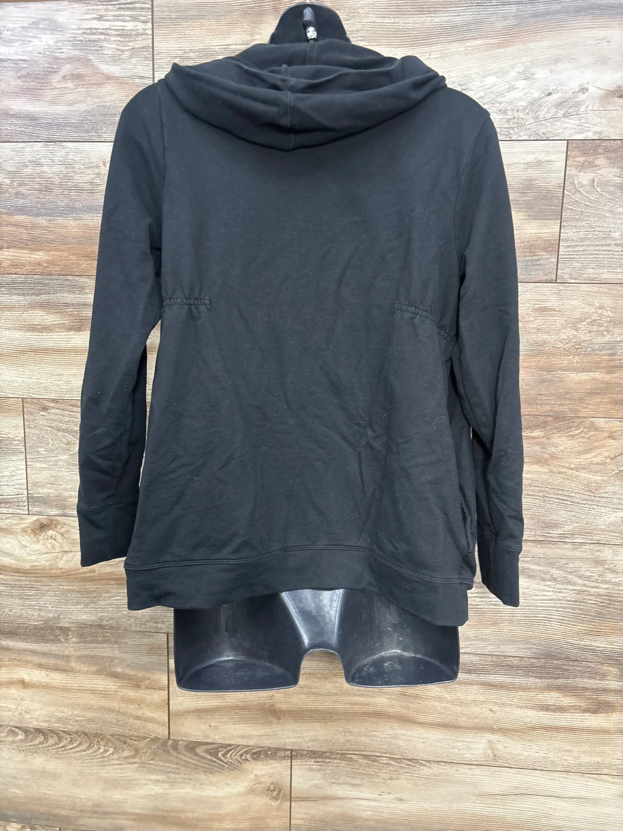 Back view of black maternity zip-up hoodie, size large, on mannequin showing hood and side ruching against wood panels