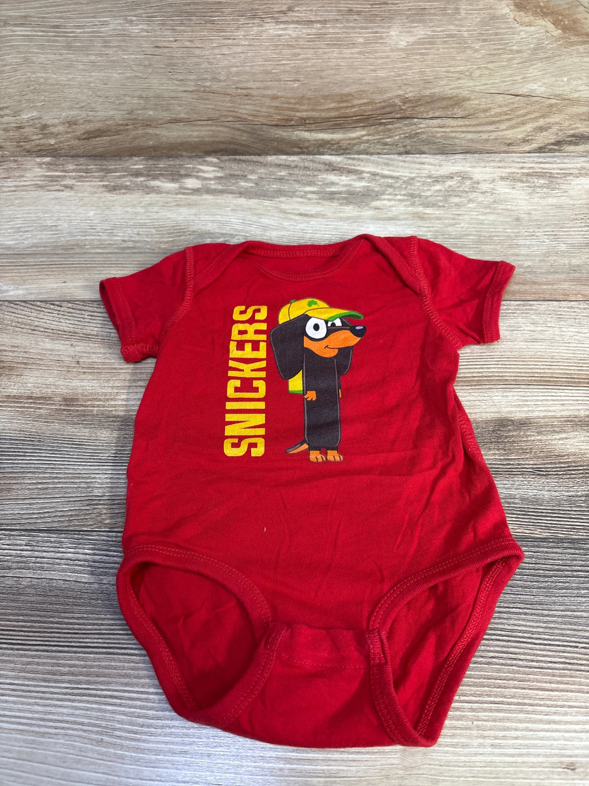 Flat lay front of red baby bodysuit showing yellow vertical SNICKERS text and cartoon dachshund graphic on wood background