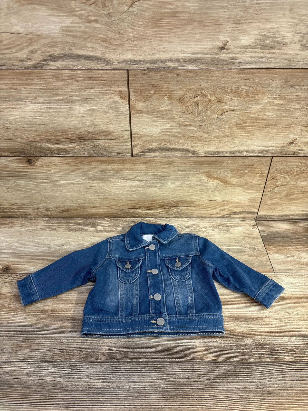 Front view of Old Navy blue denim jacket for ages 3 to 6 months with long sleeves and buttoned flap pockets on a wood-look background