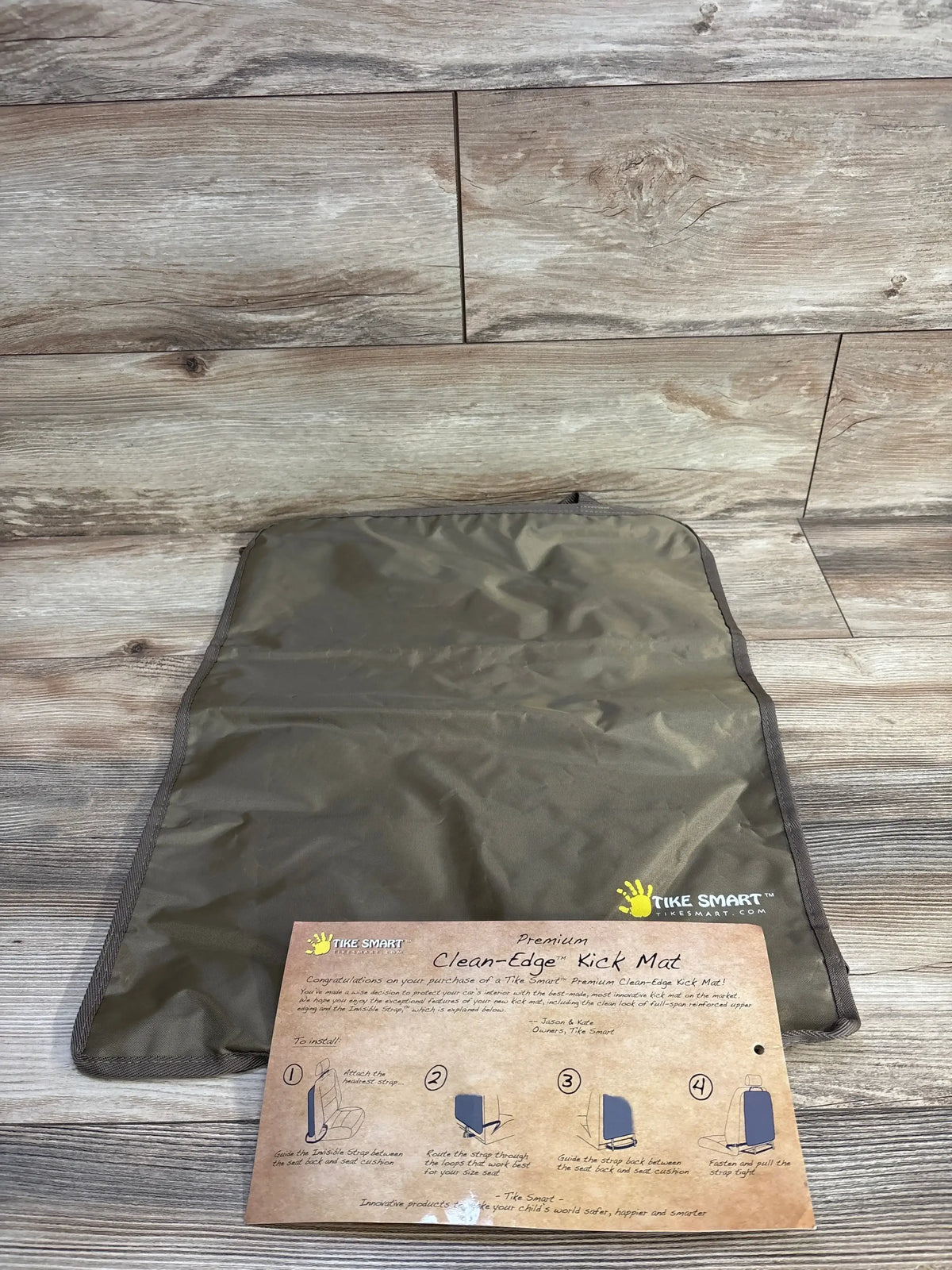 Top-down view of Tike Smart Clean-Edge Kick Mat showcasing nylon surface and logo with included instruction card on wood plank background