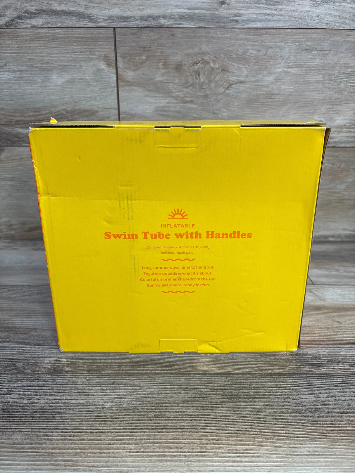Front view of Sun Squad Swim Tube with Handles box showing the orange product title and size text on a yellow package with a wood floor and wall background
