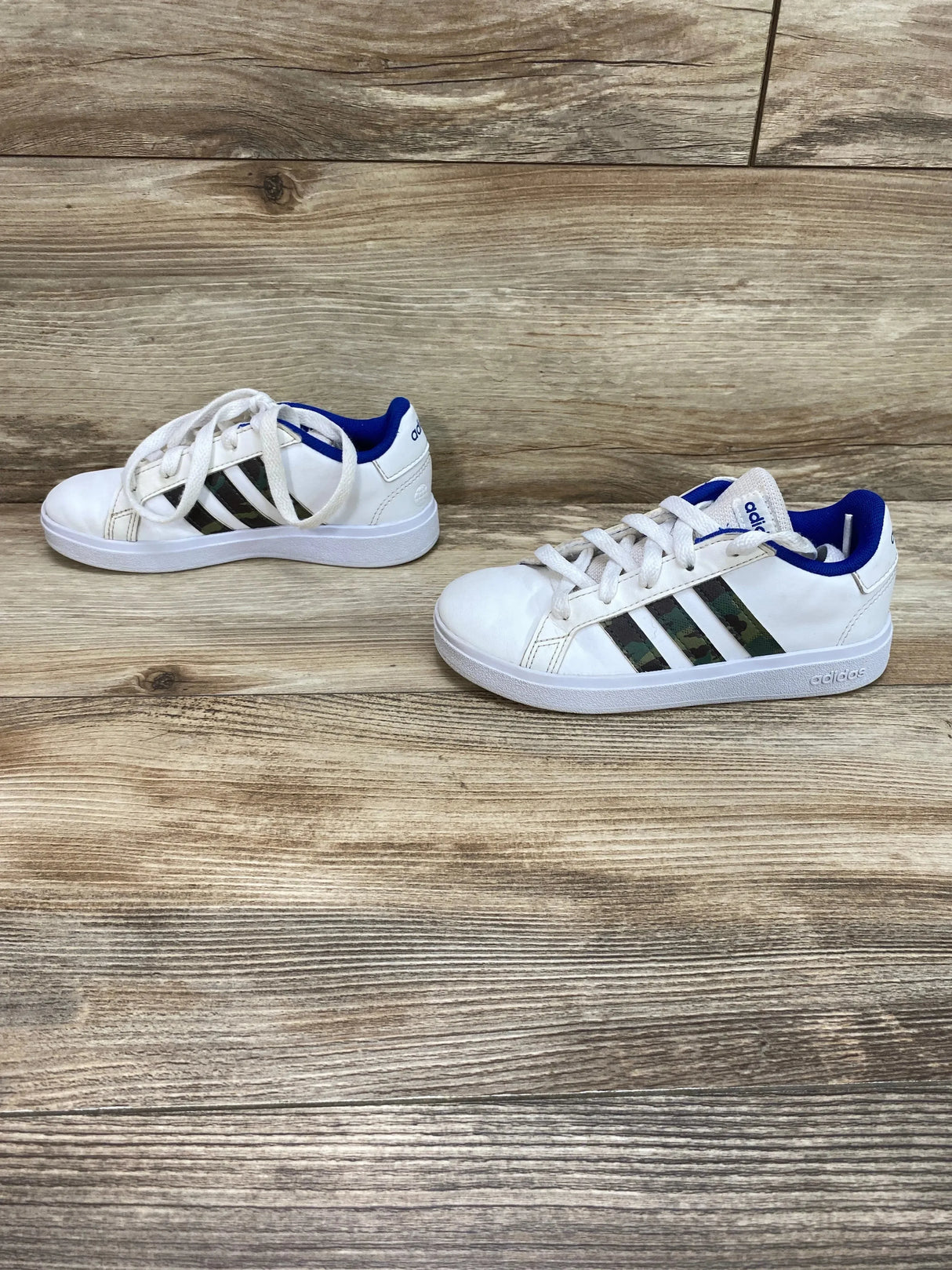 Right-side view of Adidas Grand Court Base toddler sneaker, white with camo three stripes and royal blue lining, size 12C on wood floor
