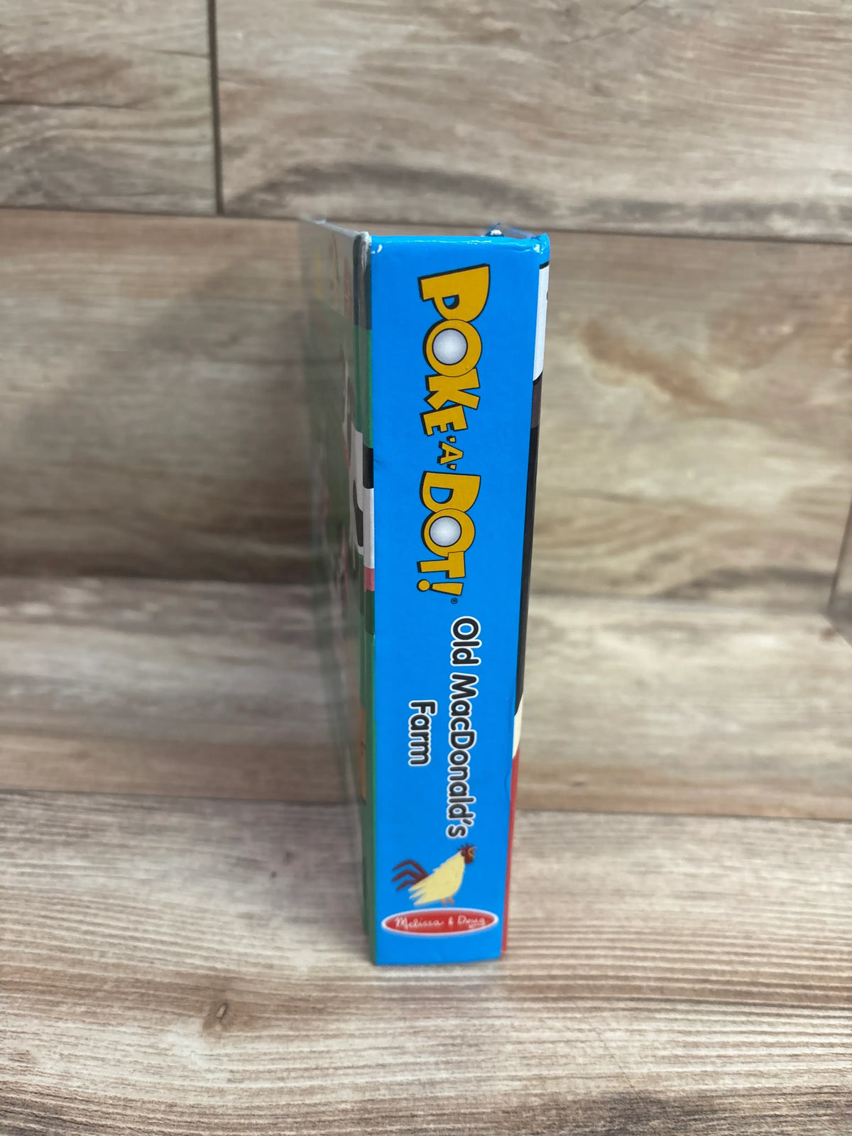 Side view of Melissa and Doug Poke-a-Dot Old MacDonald’s Farm board book showing blue spine on a wood floor