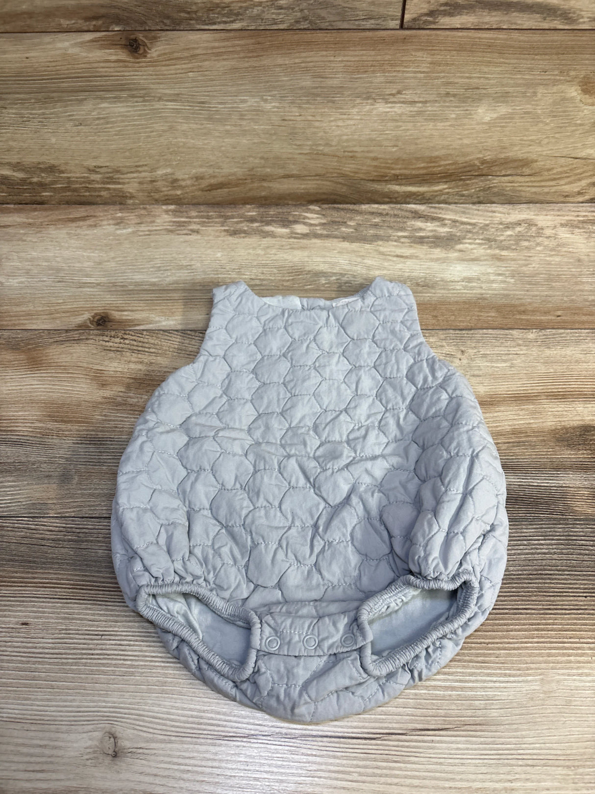 Flat lay of grey sleeveless quilted bubble romper showing scallop quilting, elastic leg openings and snap closures on wooden background