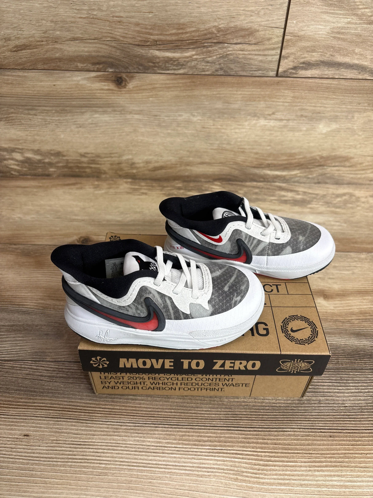 Three-quarter view of Nike Kyrie 8 TD sneakers in white and university red with grey overlay on Move to Zero box on wood plank floor