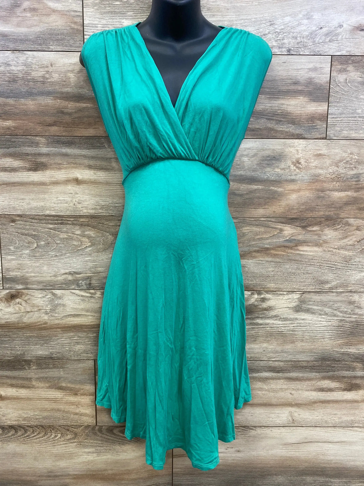 Front view of Maternal America sleeveless maternity dress, green, showing V-neck wrap bodice and empire waist on wood background