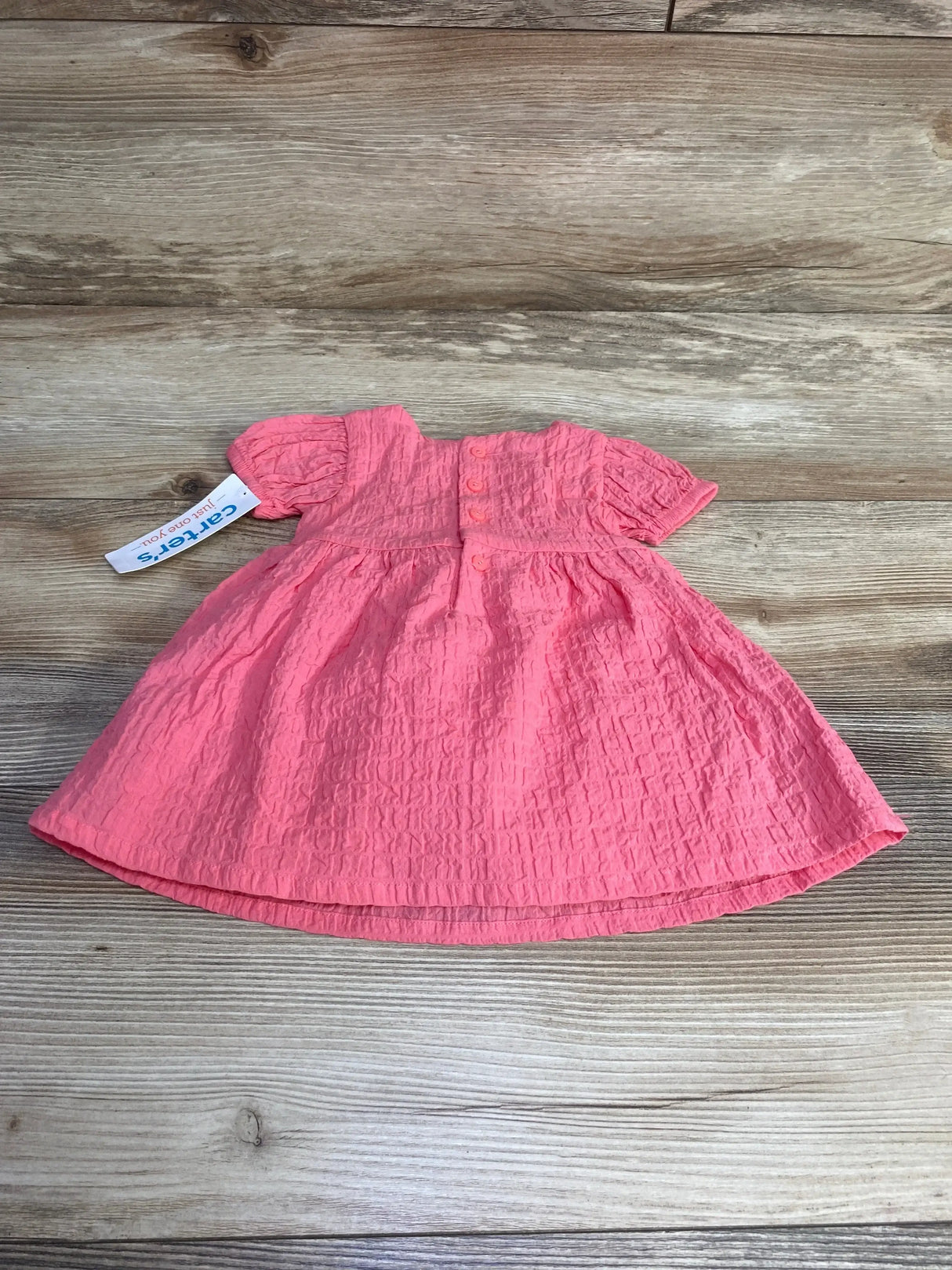 Flat lay of newborn pink textured dress showing short puff sleeves, three front buttons and gathered skirt on wood background