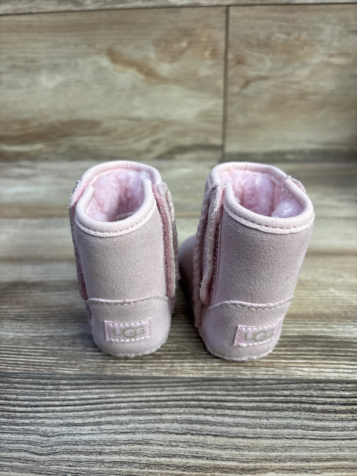 Rear view of UGG baby classic soft sole boots in pink showing suede exterior, plush pink shearling lining, UGG heel label on wood-textured background