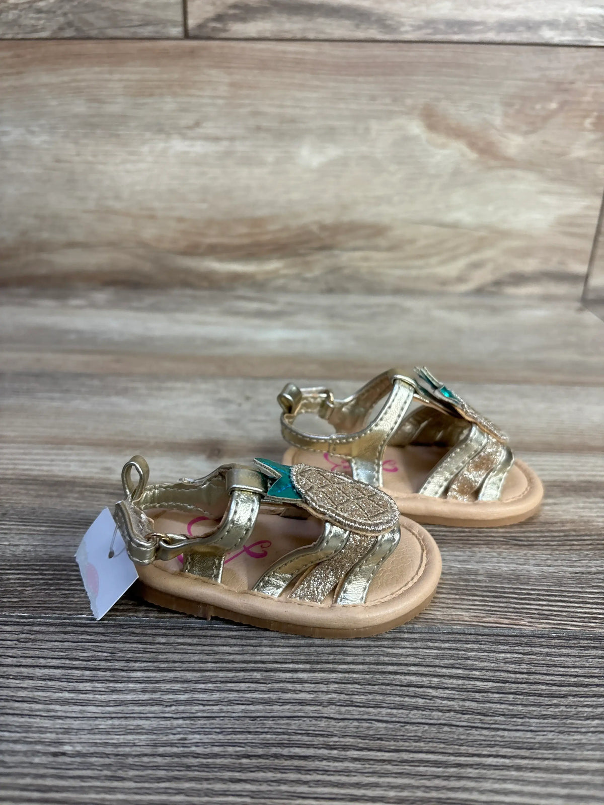 Front three-quarter view of girls pineapple sandals showcasing metallic pineapple applique and ankle straps on a wood surface
